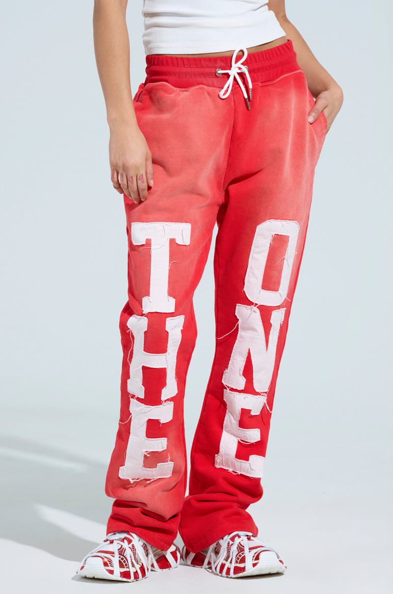 A person wears vibrant red ONLY ONE WIDE LEG PANTS featuring "THE ONE" in large white letters and an elastic waistband for comfort. These stylish pants pair perfectly with red and white sneakers, creating a bold look. 1