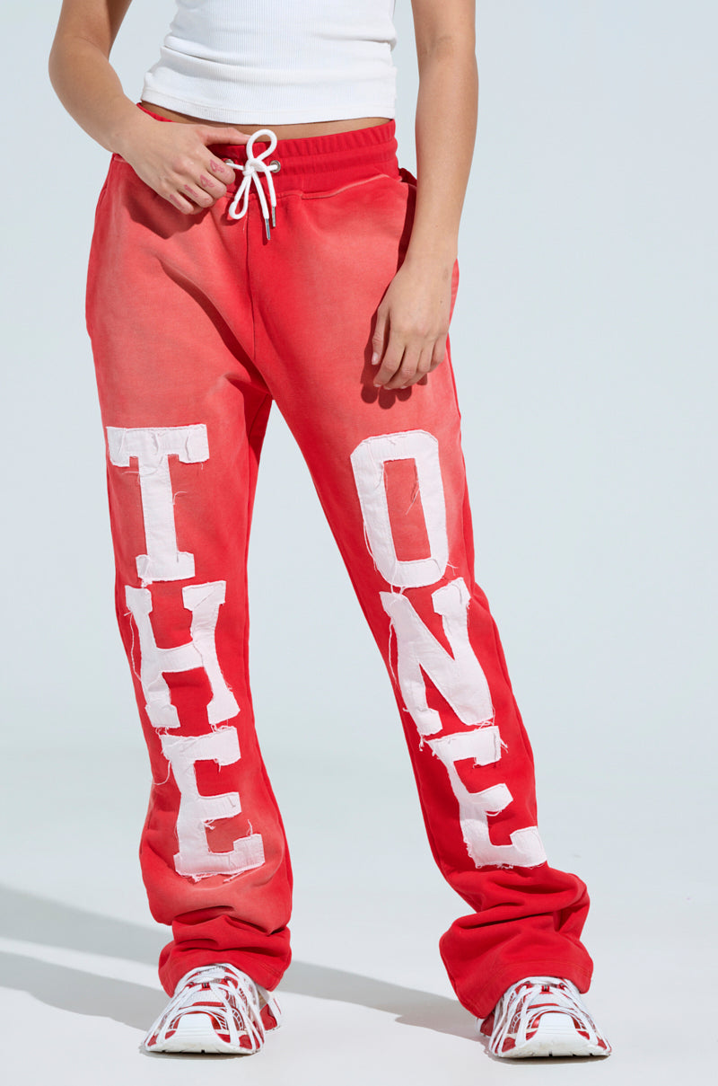 Someone is wearing red ONLY ONE WIDE LEG PANT with an elastic waistband featuring "THE" embroidered on one leg and "ONE" on the other in white patches. They're paired with a white tank top and sleek red-and-white sneakers against a plain, light-colored backdrop. 2