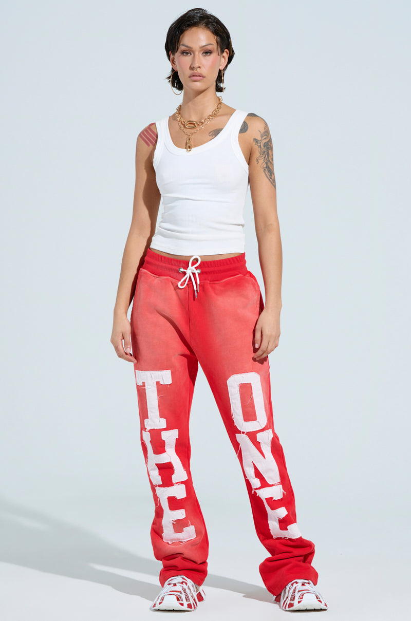 A person in a white tank top and "ONLY ONE" elastic waistband red sweatpants, featuring large white letters. They have short hair, arm and shoulder tattoos, a necklace, and white sneakers against a plain background. 6