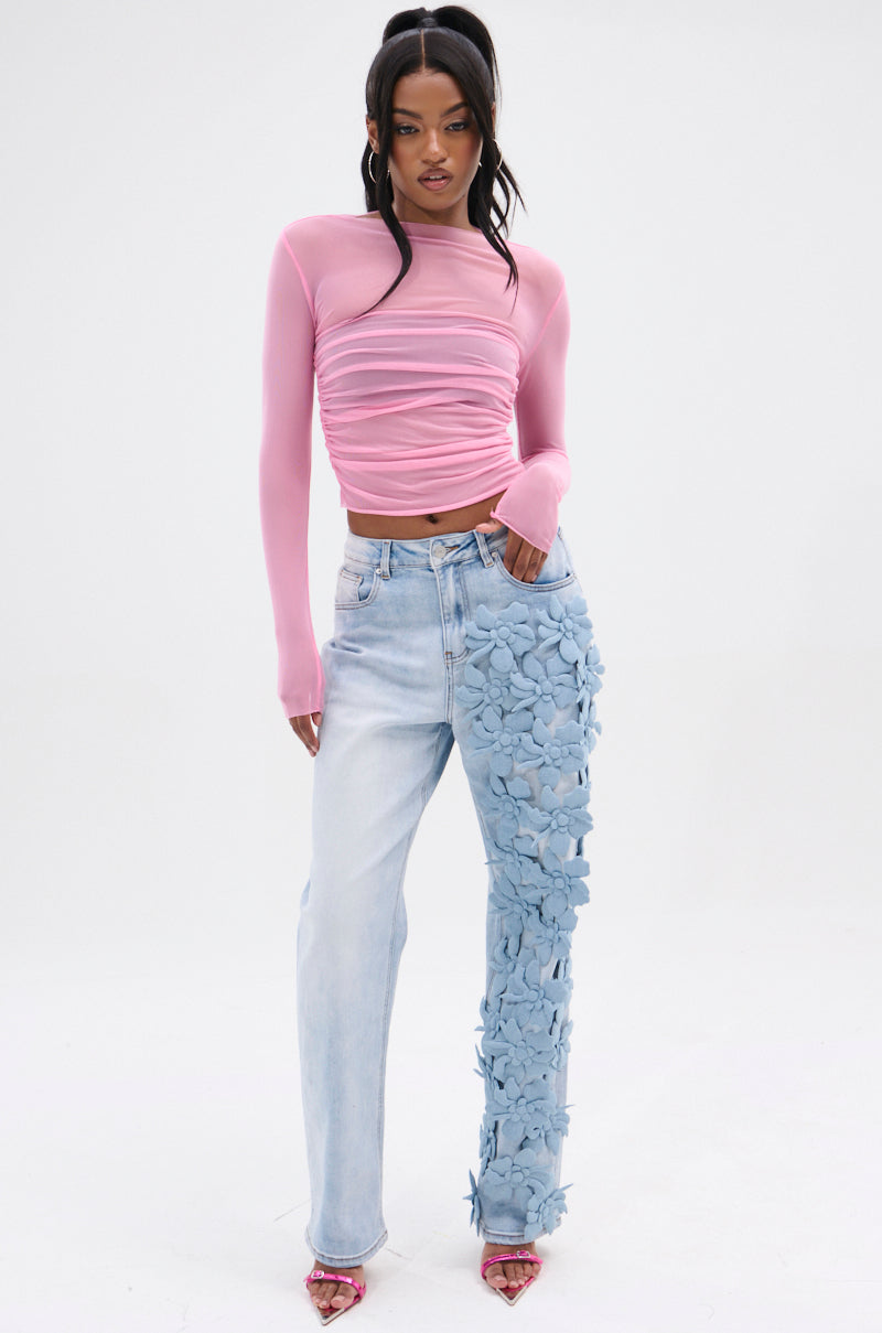A person exudes confidence in a pink, long-sleeve ruched top with high-rise FLORAL FIELDS APPLIQUE DENIM PANTS on one leg. With their dark hair styled in a high ponytail, they complete the ensemble with open-toed pink heels.