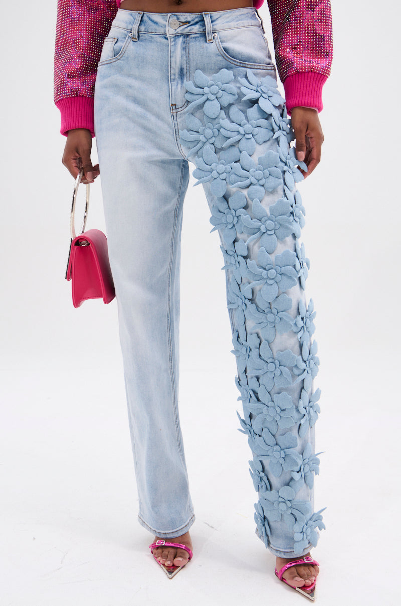 Wearing trendy high-rise FLORAL FIELDS APPLIQUE DENIM PANTS in light blue, they highlight the 3D floral design on one leg. The stylish ensemble includes a small pink handbag, a pink sweater, and matching pink heels, set against a pristine white backdrop.