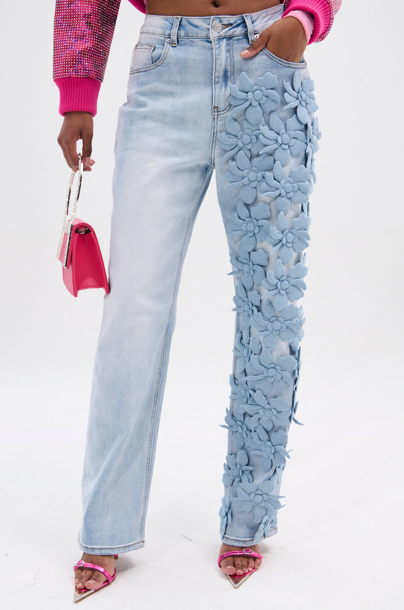 Dressed in light blue FLORAL FIELDS APPLIQUE DENIM PANTS with floral designs on one leg, a person holds a small pink handbag. The outfit features pink sandals and a sparkly pink sweater, all against a plain white backdrop.