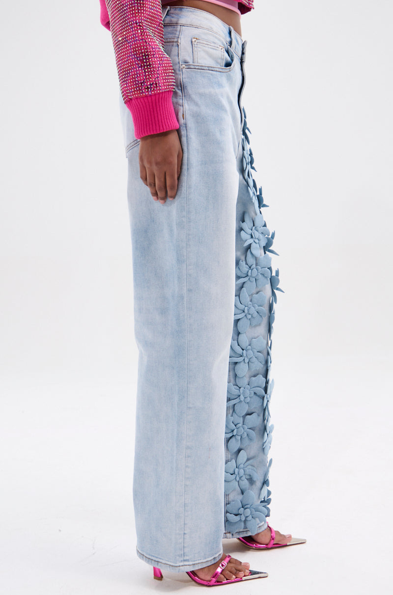 A person wears high-rise light blue denim pants featuring large Floral Fields Applique on one leg, a pink textured long-sleeve top, and pink heeled sandals, set against a plain white background.