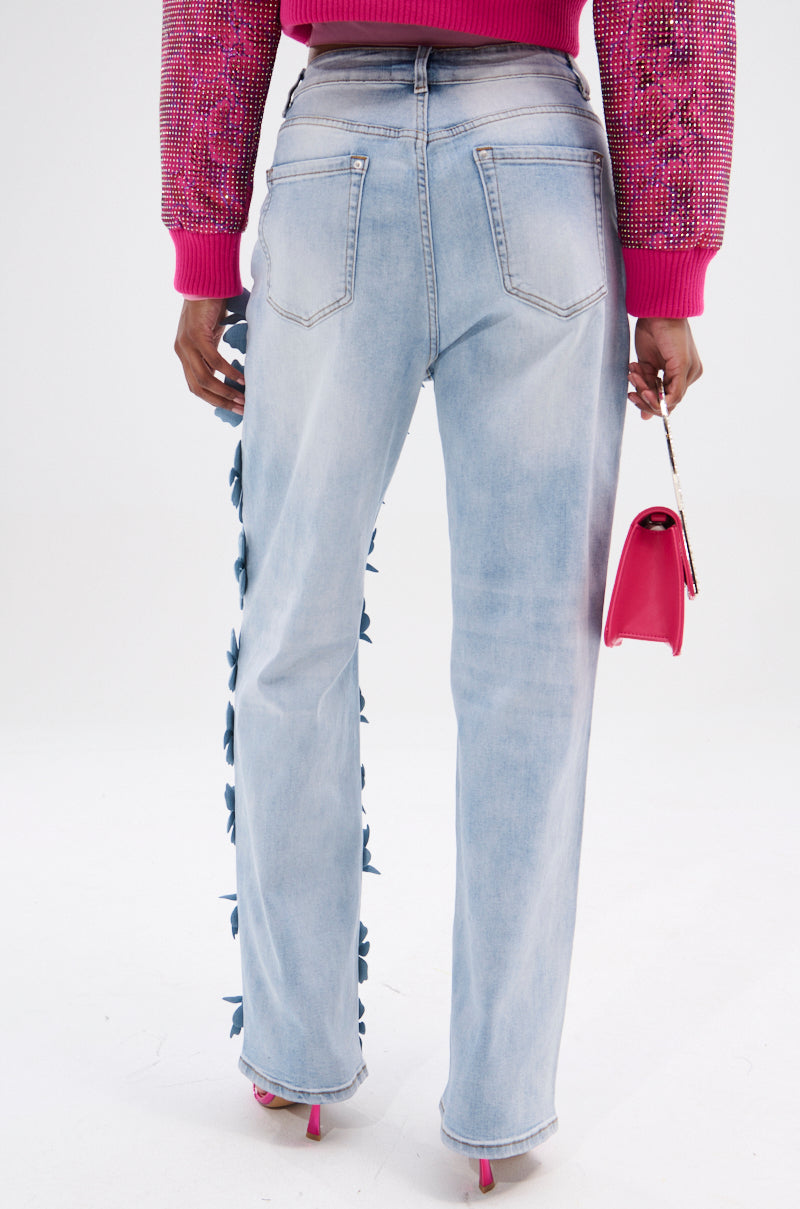 Dressed in FLORAL FIELDS APPLIQUE DENIM PANTS, featuring a high-rise fit and light blue color with floral embellishments on one leg, a person pairs them with a pink sweater and matching small handbag while posing gracefully against a plain white background.