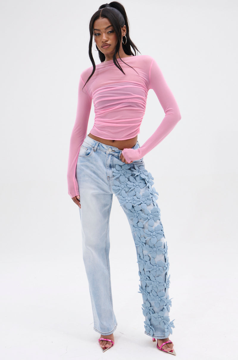 A person stands against a plain background wearing a pink long-sleeve top, FLORAL FIELDS APPLIQUE DENIM PANTS, featuring a floral design on one leg. They have long hair styled into two ponytails and are paired with pink heels.