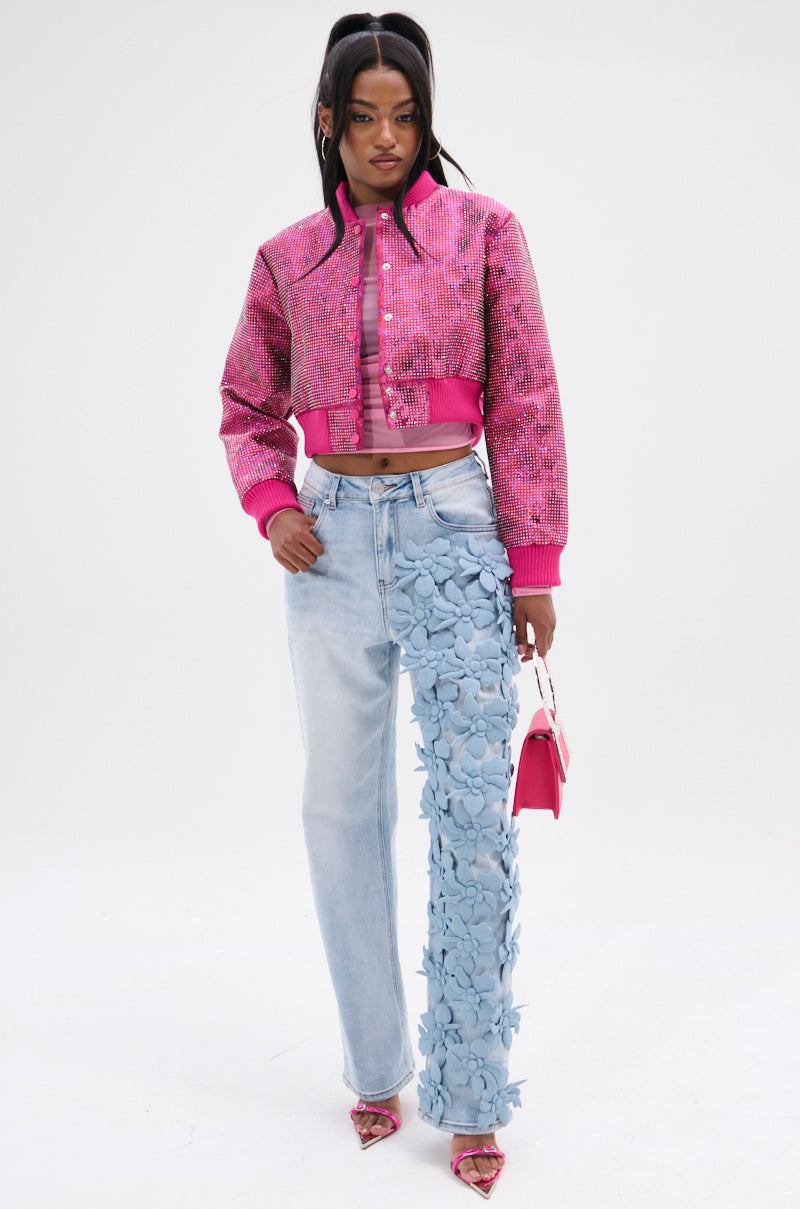 Against a plain backdrop, someone models a textured pink jacket and the FLORAL FIELDS APPLIQUE DENIM PANTS with flower designs on one leg. They accessorize with pink shoes, a small pink handbag, and style their hair in a high ponytail for an effortlessly chic appearance.