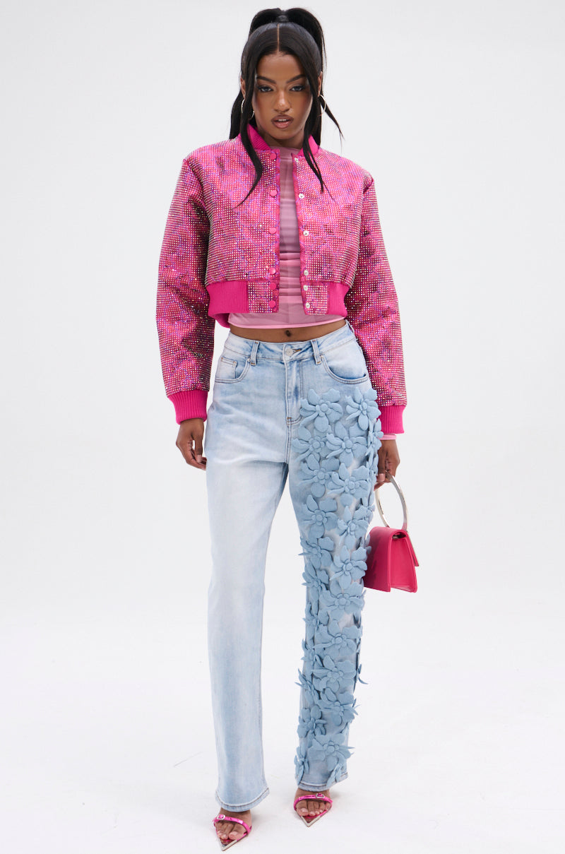 A person in a pink textured jacket, sheer pink top, and FLORAL FIELDS APPLIQUE DENIM PANTS is holding a small pink bag and wearing matching heels against a white backdrop.