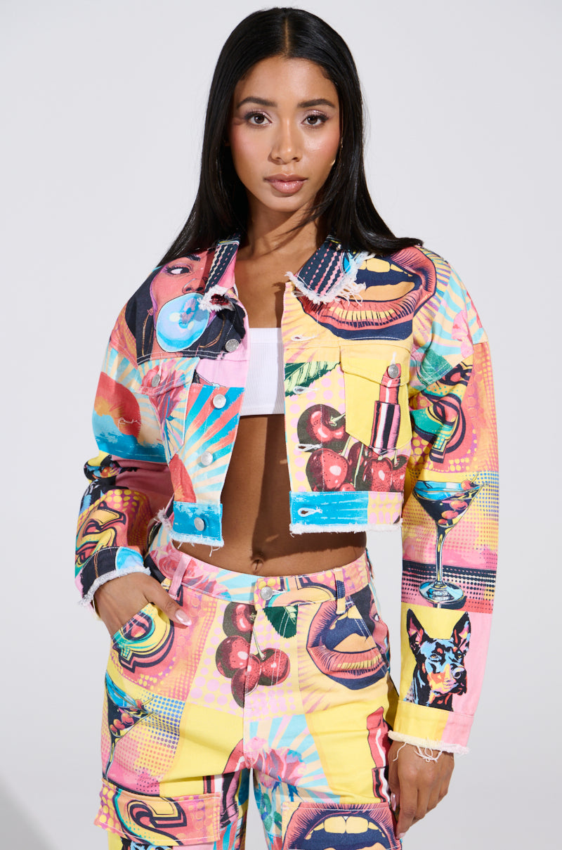 A woman with long dark hair models the ELECTRIC PRINTED DENIM JACKET and matching pants, featuring bold technicolor prints of lips, a cat, and a martini glass. She wears a white top underneath the colorful set against a plain backdrop.
