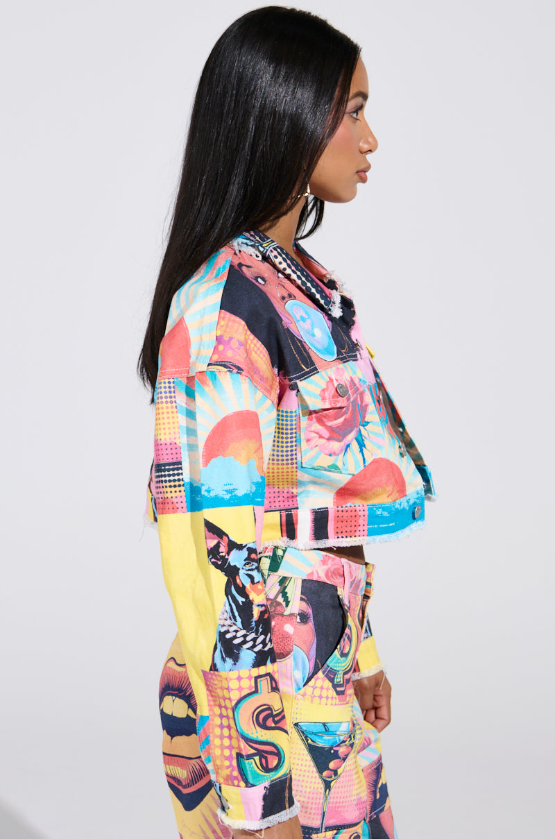 A woman with long black hair is shown in profile, wearing an ELECTRIC PRINTED DENIM JACKET adorned with colorful pop art-inspired abstract shapes and faces. The plain white background highlights her vivid style.