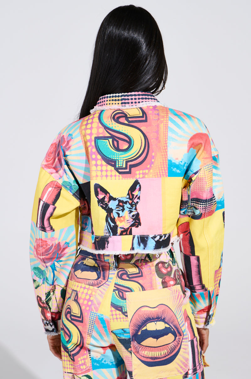 A person with long black hair wears the ELECTRIC PRINTED DENIM JACKET, showcasing a pop art-inspired look with technicolor graphics of lips, a dog, roses, and vibrant patterns in yellow, pink, and blue on a plain white background.
