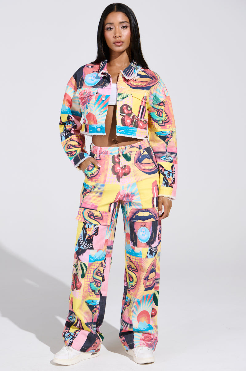 A woman exudes confidence in an ELECTRIC PRINTED DENIM JACKET, a colorful pop art-inspired two-piece with cropped jacket and wide-leg pants featuring vibrant technicolor graphics. She completes the look with white sneakers and straight long hair.