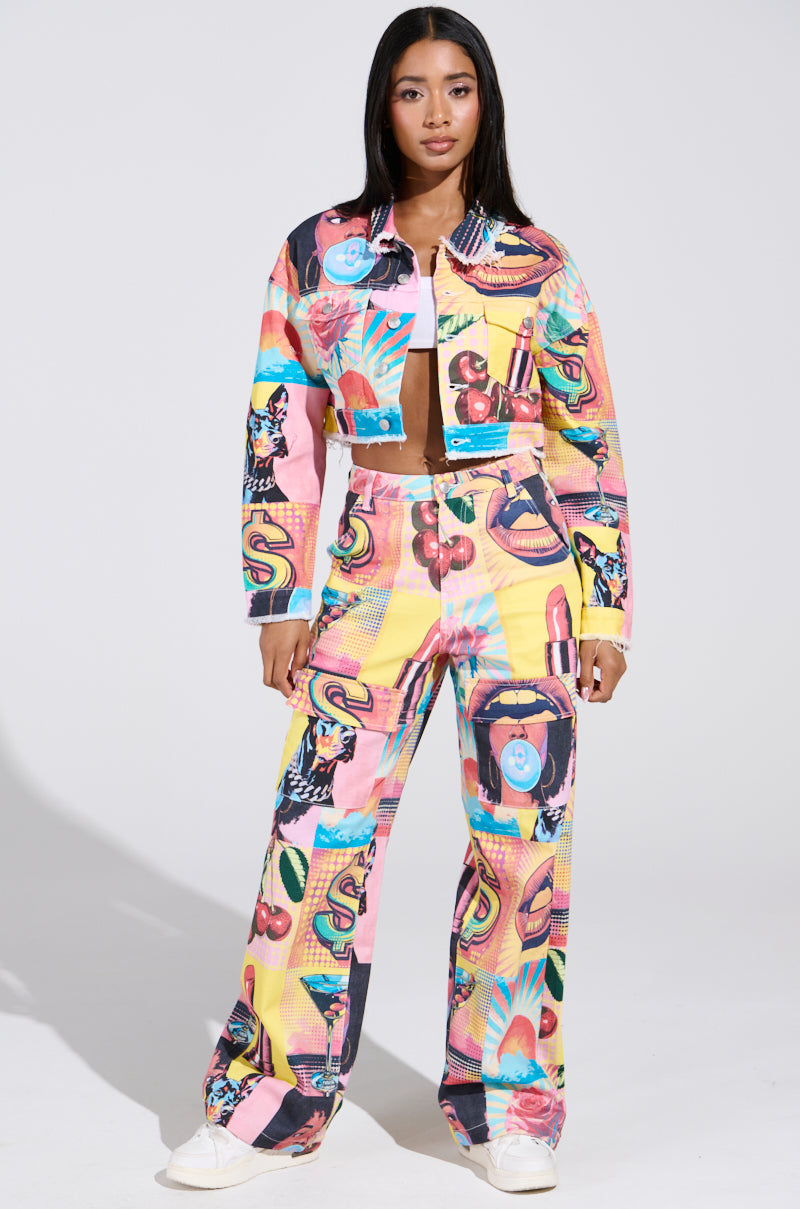 A woman stands confidently in an ELECTRIC PRINTED DENIM JACKET, featuring a pop art-inspired mix of patterns like flowers and fruits. Her long dark hair cascades as she relaxes in white sneakers against a plain backdrop, embodying vibrant energy.