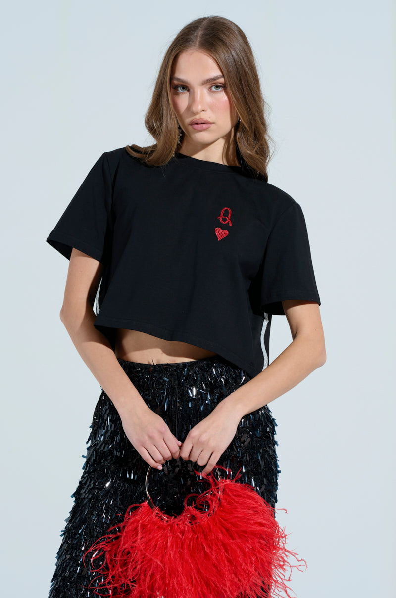 A woman in the LUCKY WINNER T-SHIRT, featuring a queen of hearts graphic, pairs it with a sequined black skirt and holds a fluffy red bag. Her long brown hair cascades down as she poses confidently against a light background.