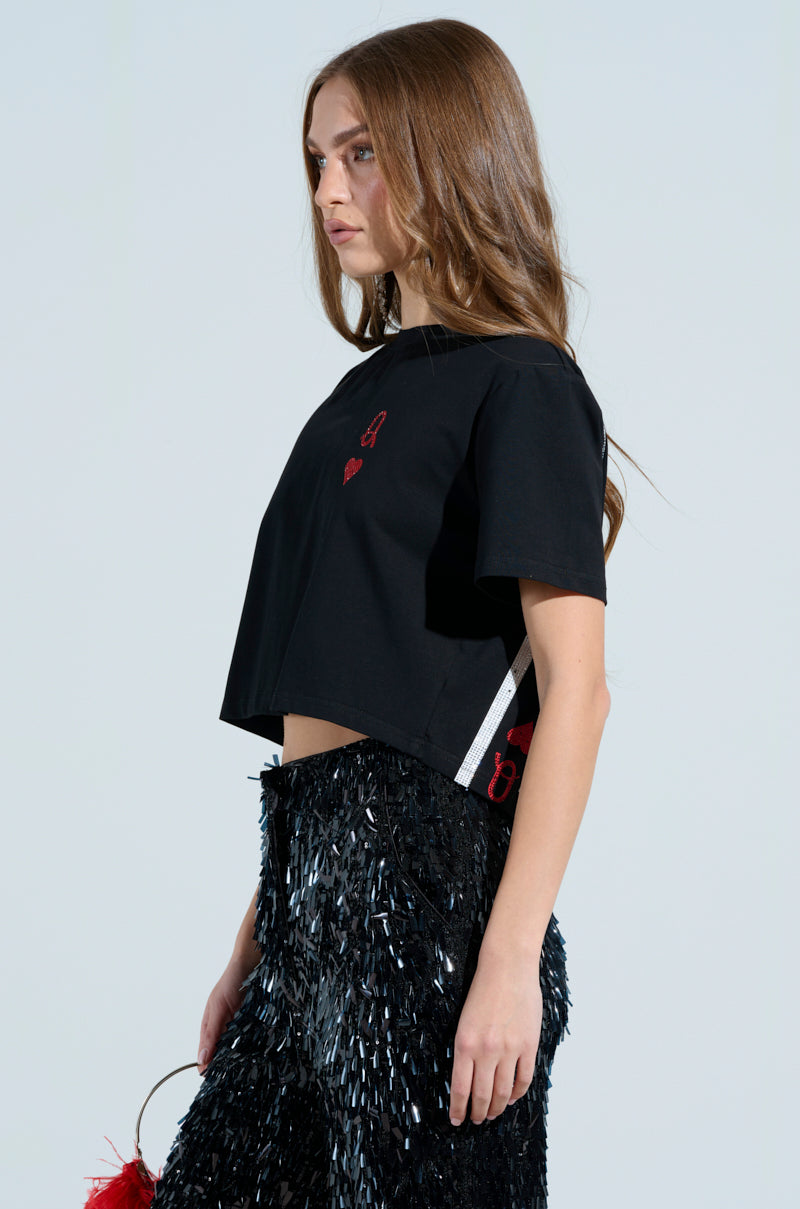 A woman with long brown hair wears the LUCKY WINNER T-SHIRT and a black skirt with rhinestone-embellished fringes. She holds a small round red bag and stands against a plain light background, looking to the side.