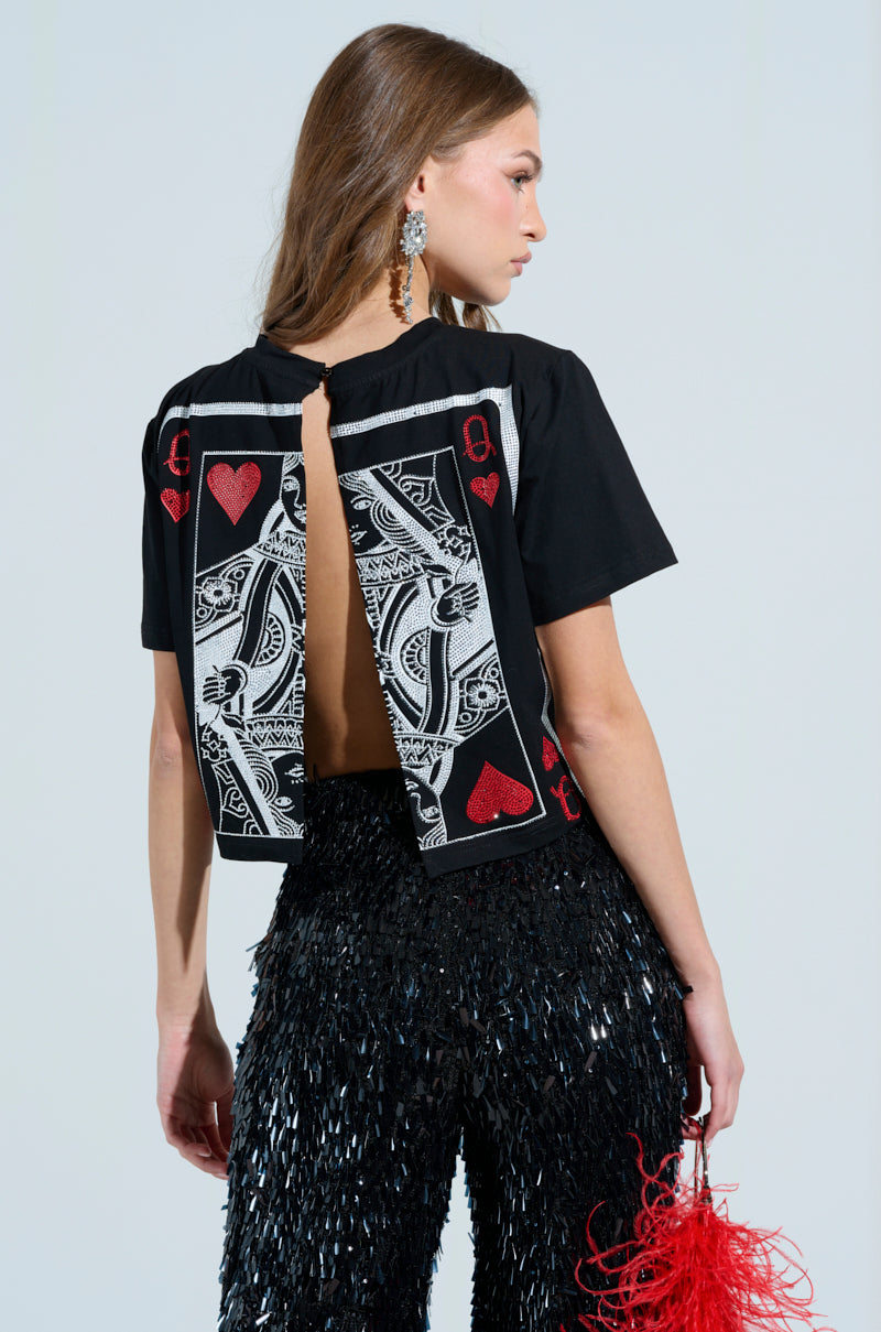 A woman with long brown hair wears the LUCKY WINNER T-SHIRT, featuring a queen of hearts graphic on the back, paired with shimmering black sequin pants. She holds a red feathery accessory in her right hand, and her look is completed with large earrings and rhinestone details.