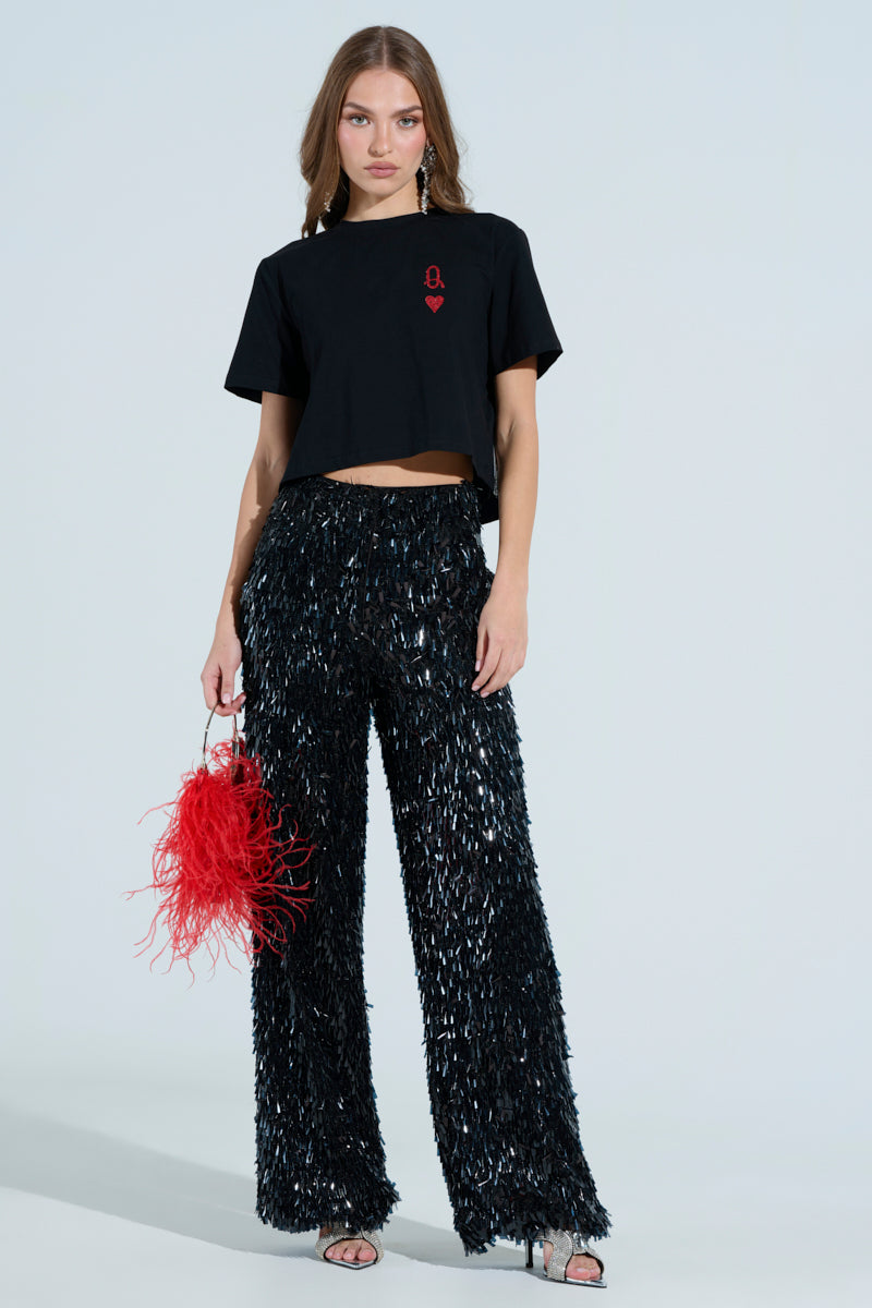 A woman stands confidently wearing a black "LUCKY WINNER" cropped t-shirt, shimmering black sequin pants, and rhinestone-embellished black high-heeled sandals. She holds a small red feathered handbag against a plain white background.