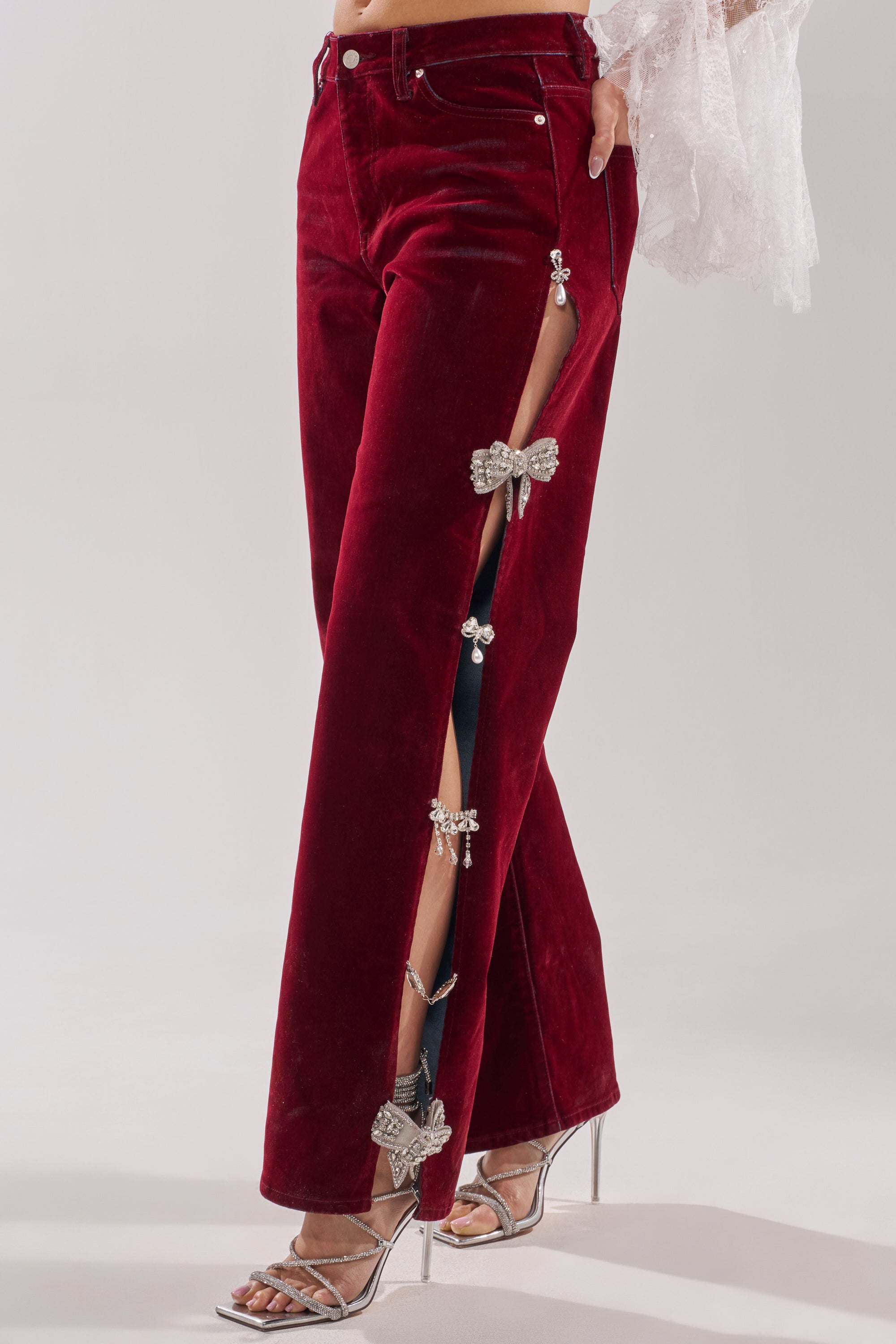 Wearing the COVERED IN DIAMONDS RELAXED VELVET DENIM in burgundy with rhinestone bow accents and side slits, styled with silver high-heeled sandals and a sheer white sleeve. 0