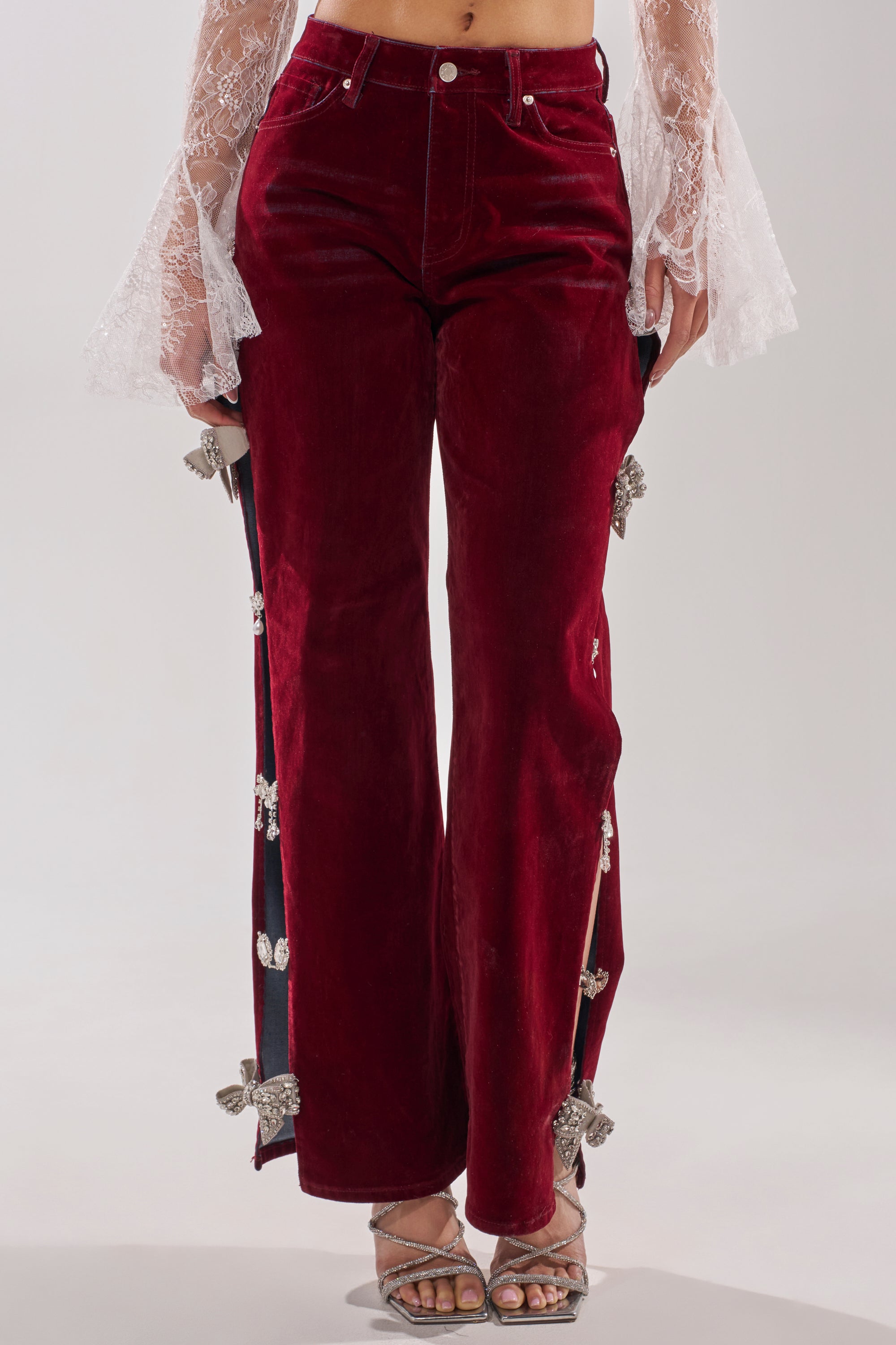 Someone stands against a plain background wearing the COVERED IN DIAMONDS RELAXED VELVET DENIM—high-waisted, straight-leg velvet pants with rhinestone bow accents—paired with a sheer white lace top and silver sandals. 1