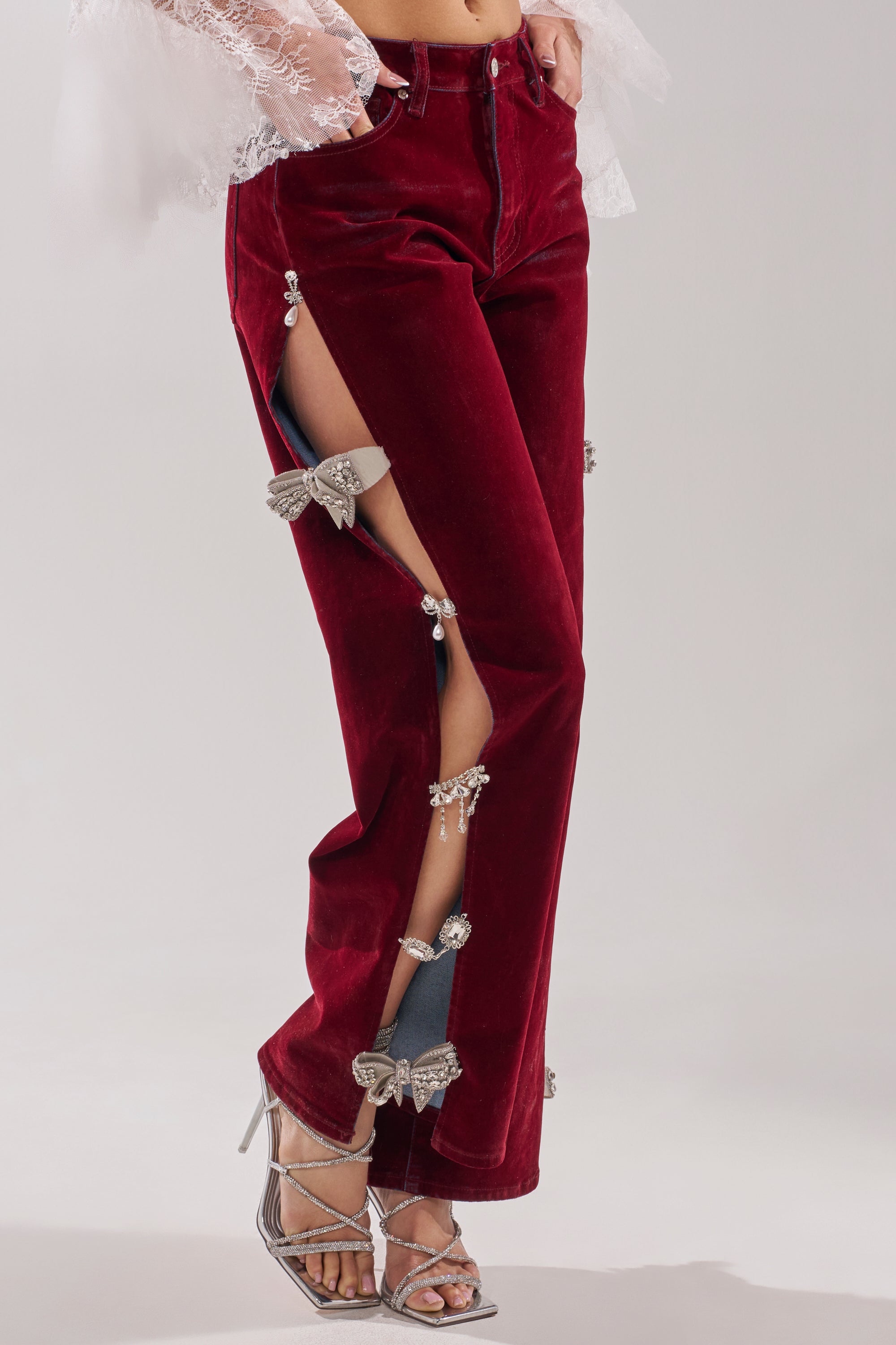 Wearing COVERED IN DIAMONDS RELAXED VELVET DENIM with open side seams and rhinestone bows, the pants reveal glimpses of the legs through sparkling details. Silver strappy heels complete the look. 2
