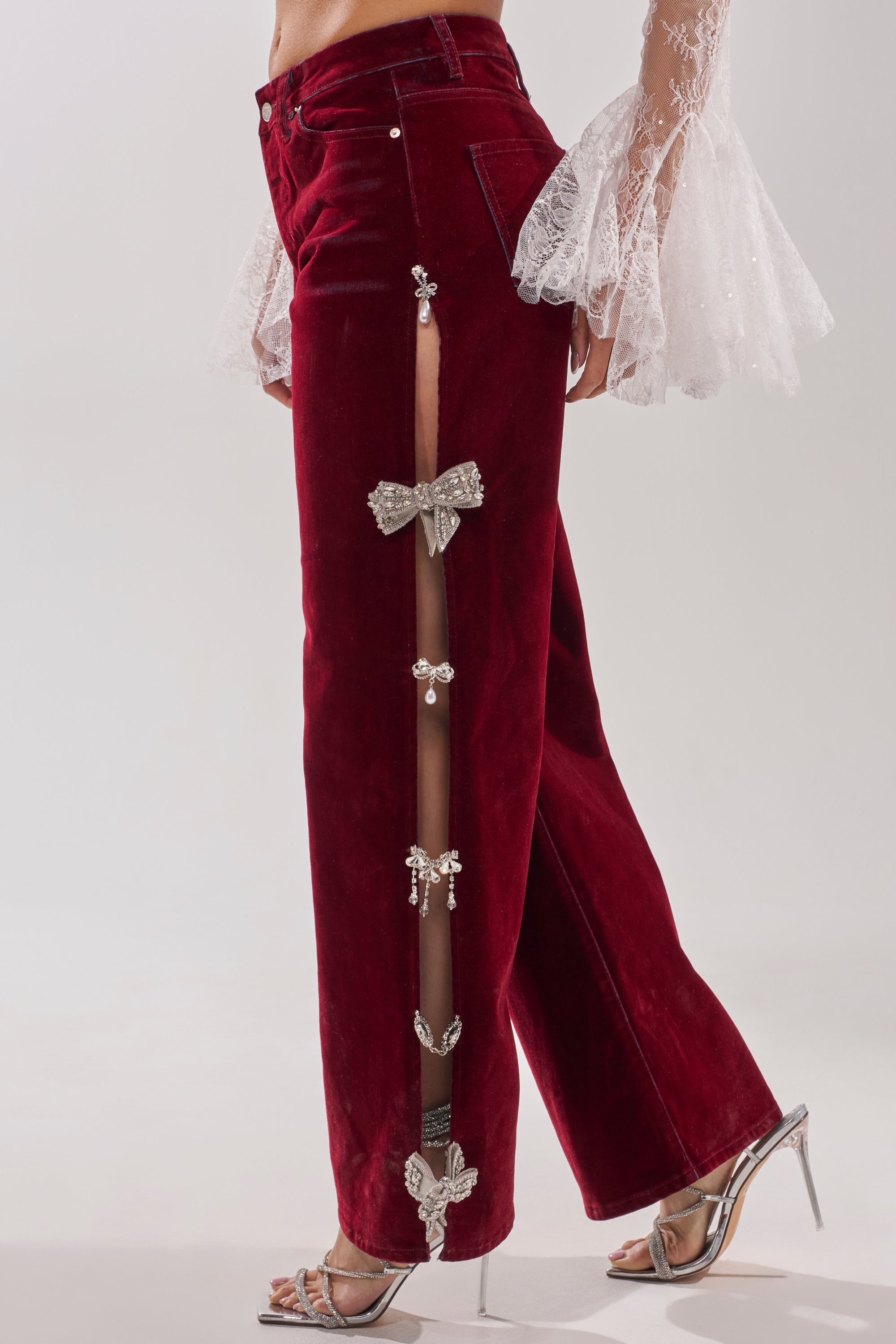 Someone wears the COVERED IN DIAMONDS RELAXED VELVET DENIM with side slits and rhinestone bow accents, styled with silver high-heeled sandals and a sheer lace sleeve top. 3