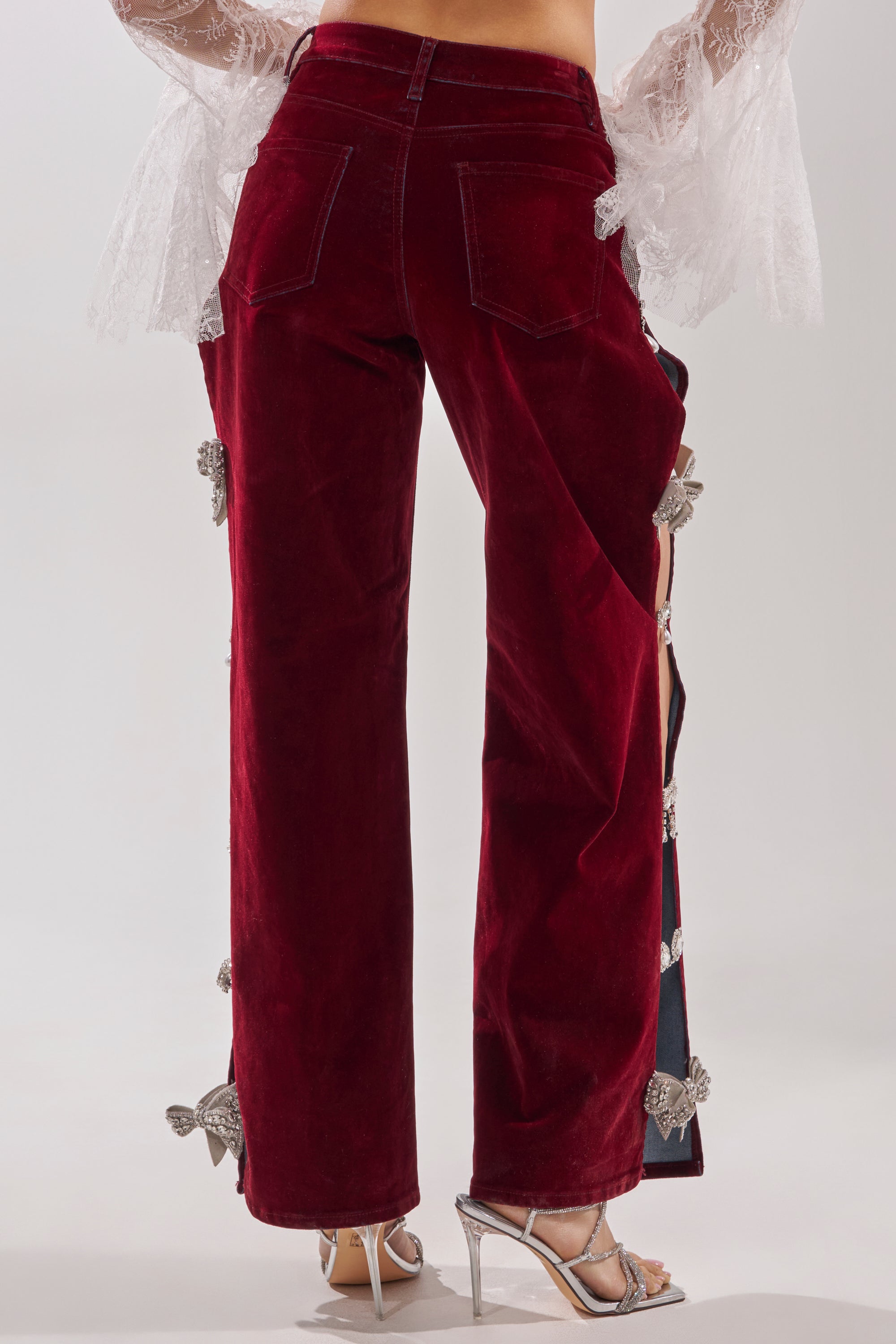 A person is shown from the back wearing COVERED IN DIAMONDS RELAXED VELVET DENIM—high-rise, straight-leg, deep red velvet pants with rhinestone bow details—paired with a sheer white lace top and silver high-heeled sandals. 4