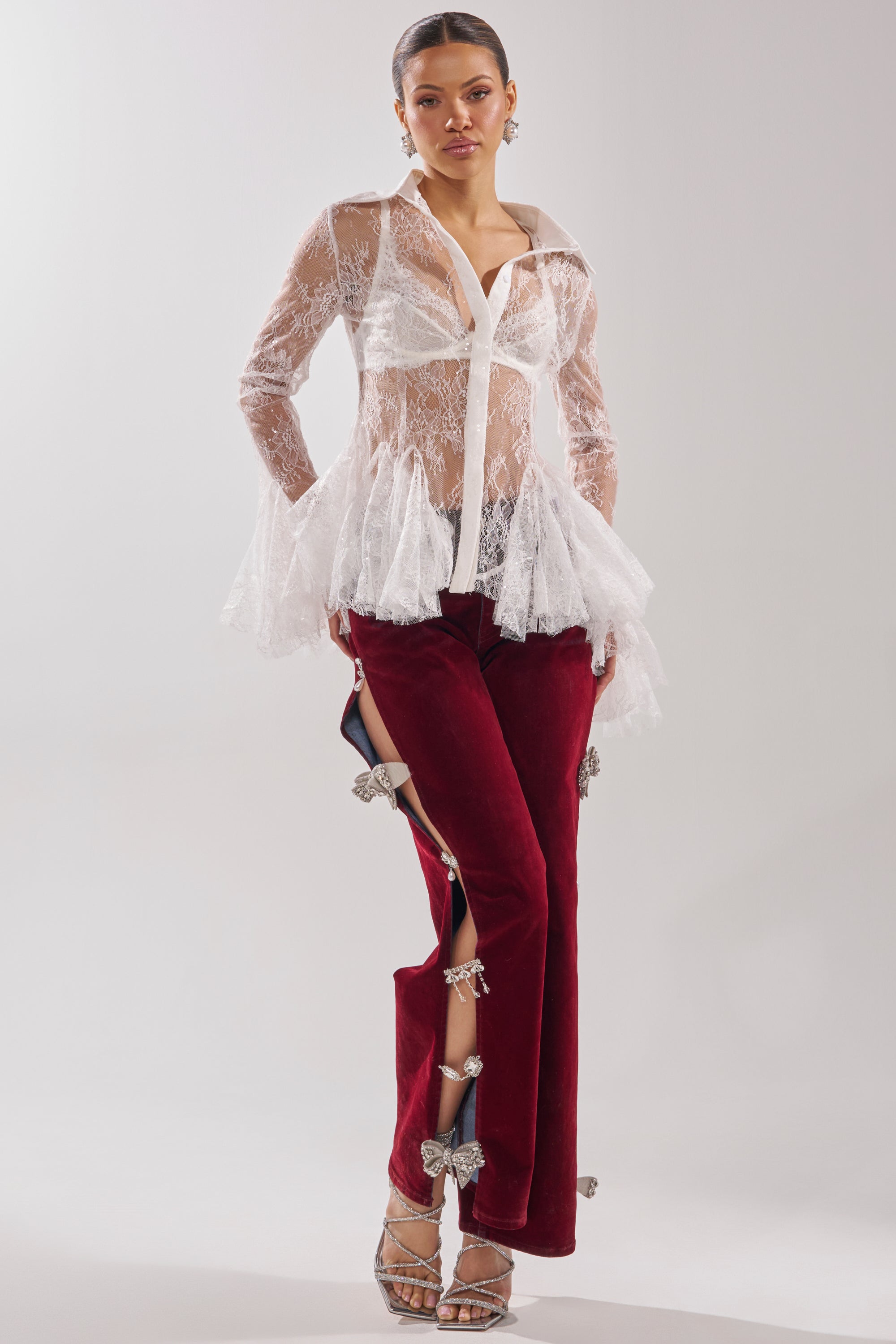 A woman poses in a sheer white lace blouse and COVERED IN DIAMONDS RELAXED VELVET DENIM—high-rise straight-leg red velvet pants with open slits, silver butterfly clasps, rhinestone bow accents—styled with strappy heels and hoop earrings. 6