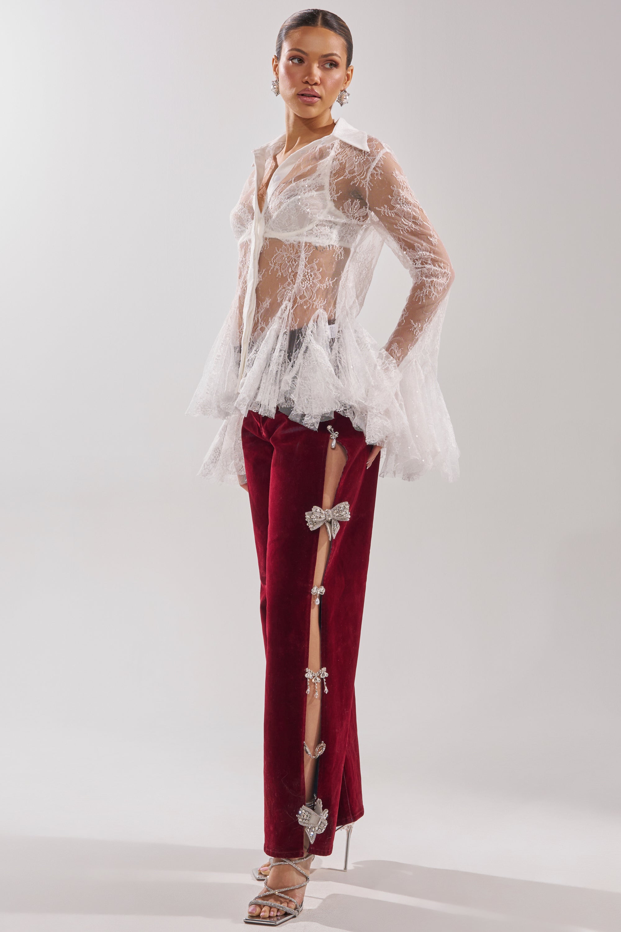 A woman poses in the COVERED IN DIAMONDS RELAXED VELVET DENIM, featuring burgundy velvet and rhinestone bow details along the side, styled with a sheer white lace blouse and silver strappy high heels. 7