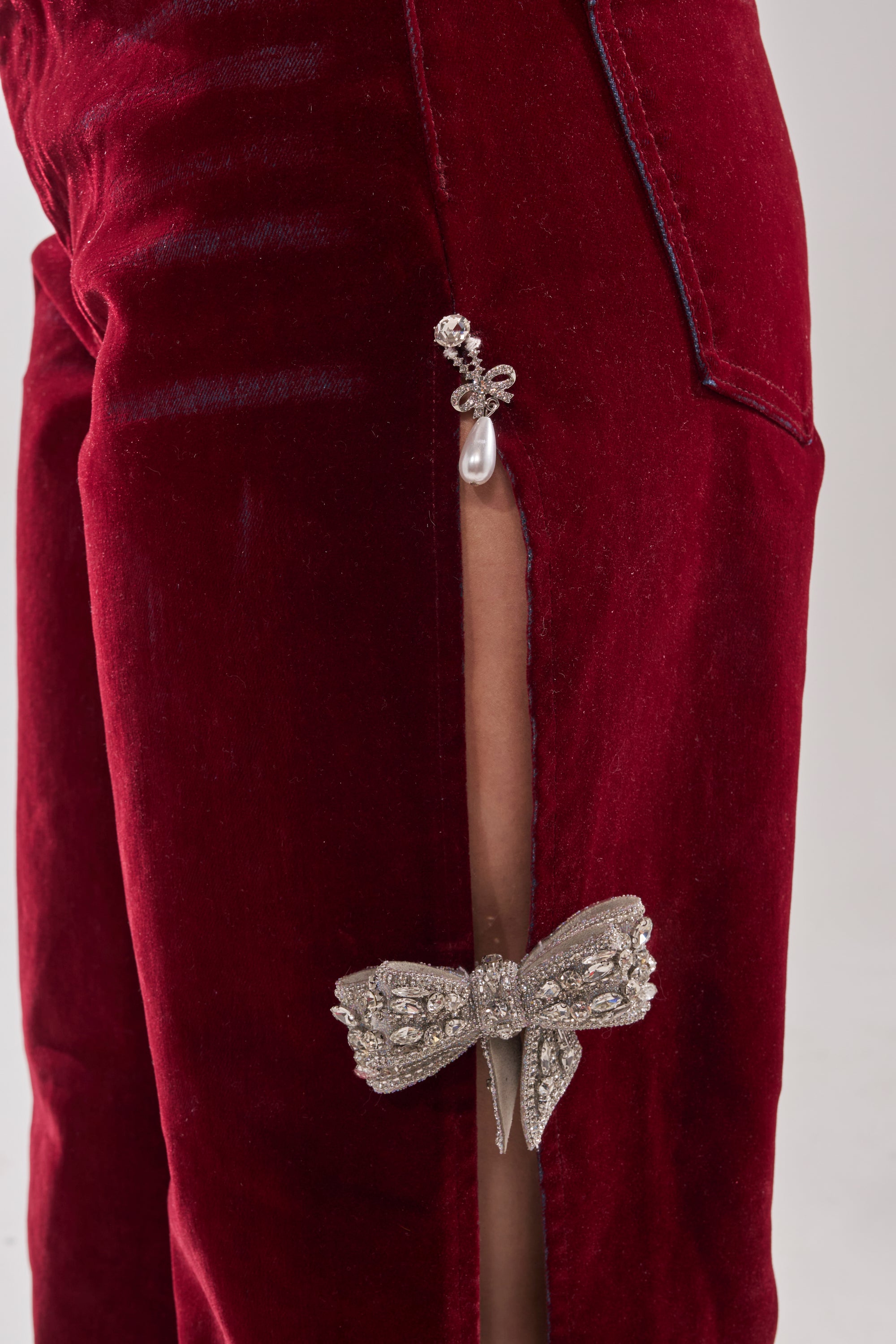 Close-up of the COVERED IN DIAMONDS RELAXED VELVET DENIM: burgundy straight-leg velvet pants with a thigh-high slit, pearl and jeweled clasp, plus a sparkling rhinestone bow near the knee. 8