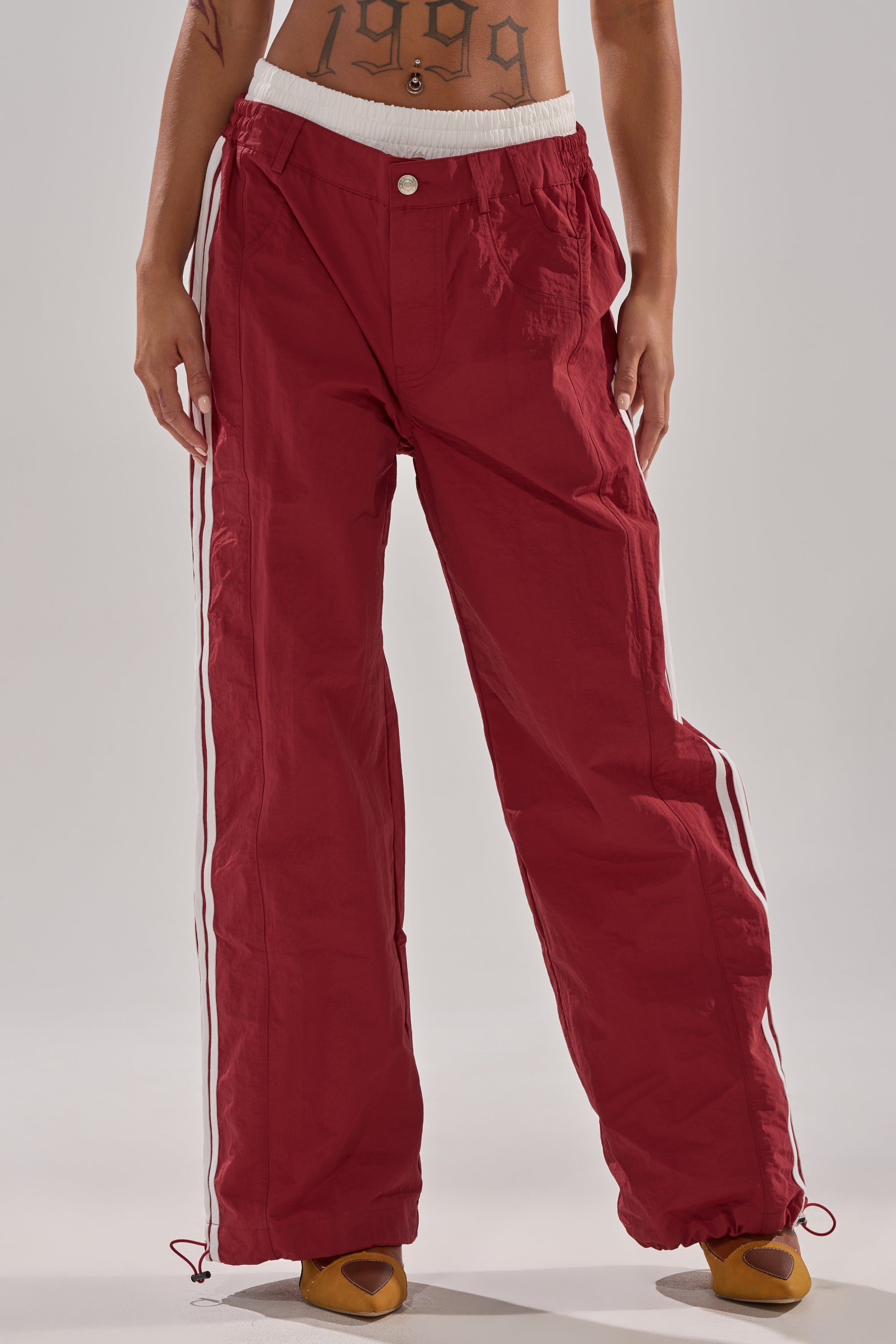 A person stands with hands by their sides, wearing the high-rise DENNIS TRACK PANT IN BURGUNDY featuring athletic stripes and a drawstring hem; the fit reveals their midriff and accentuates a tattoo across the lower abdomen. 1