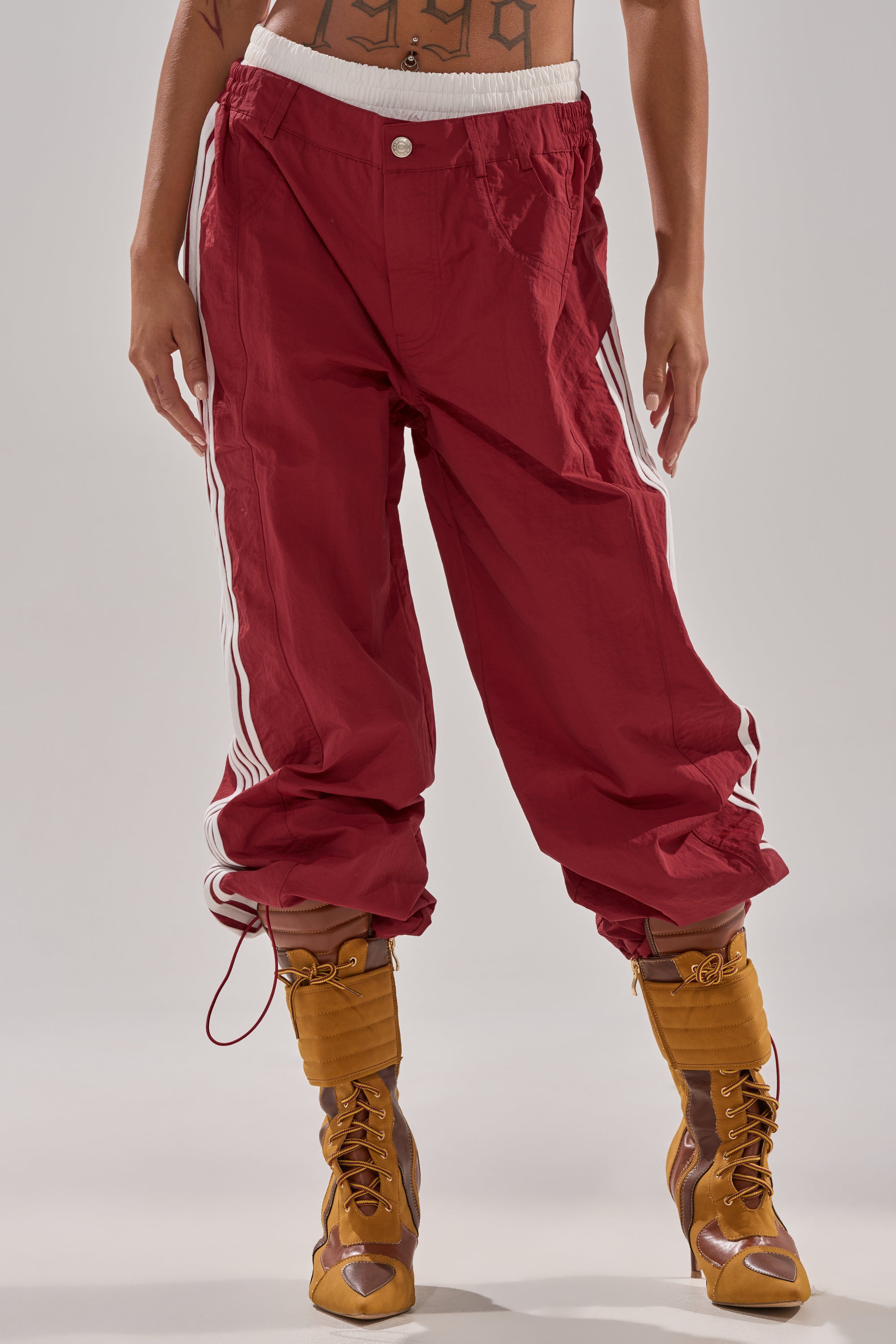 Lower torso and legs shown wearing DENNIS TRACK PANT IN BURGUNDY, a loose high-rise fit with white side stripes, styled with brown lace-up heeled boots. The background is plain and light-colored. 2