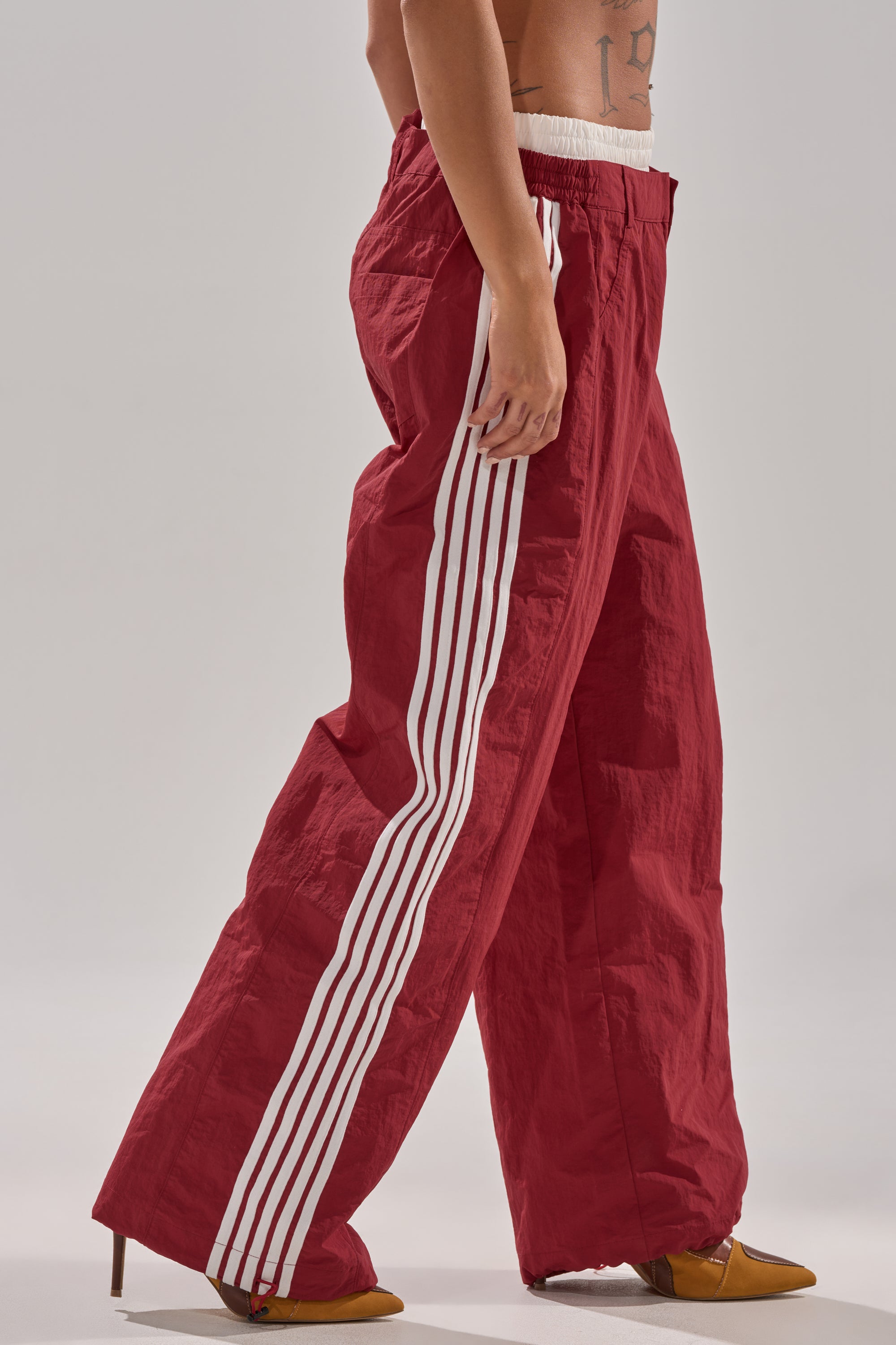 A person wears high-rise fit DENNIS TRACK PANT IN BURGUNDY with athletic stripes, paired with pointed mustard yellow heels. The cropped photo highlights the lower body against a plain background. 0