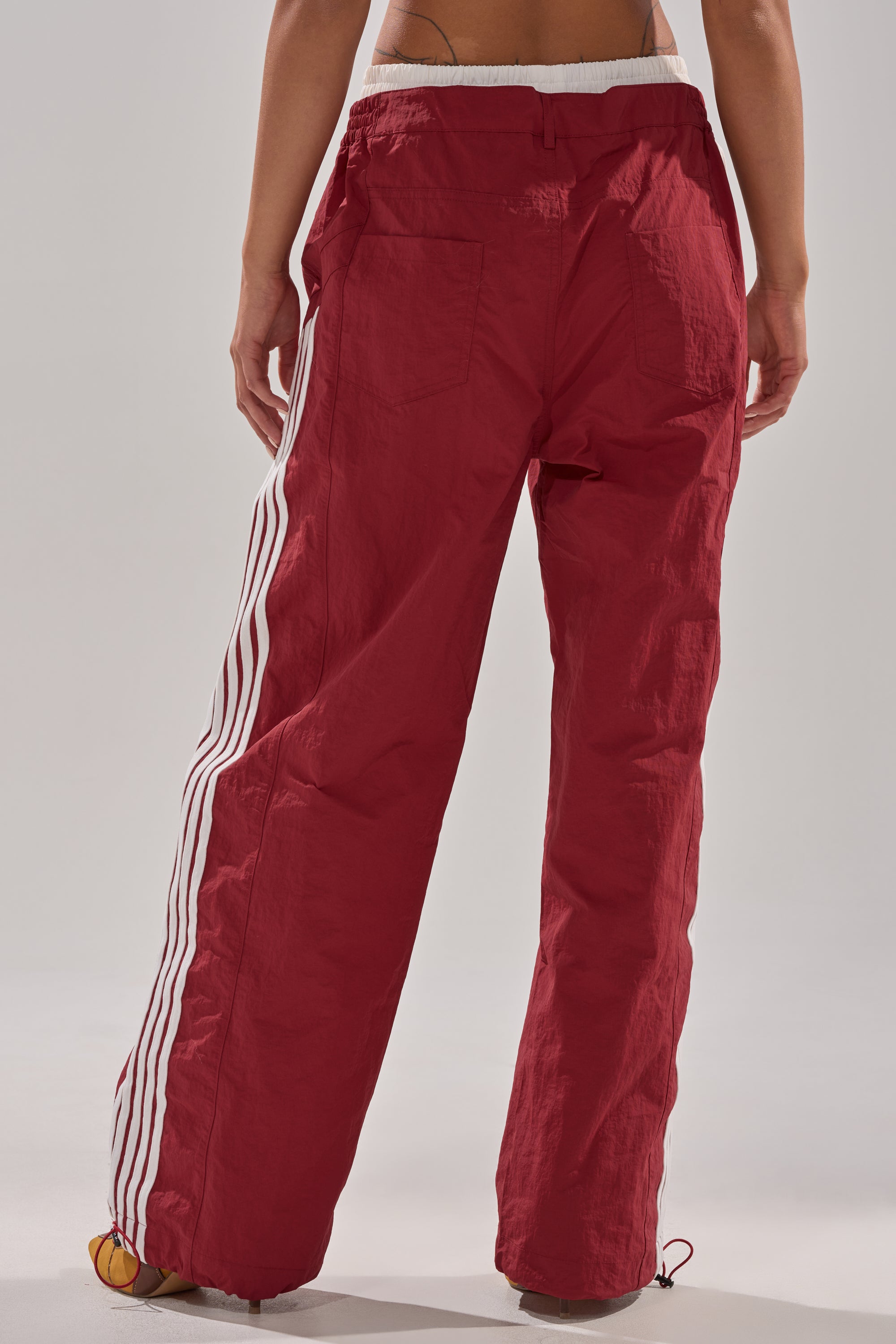 A person is seen from behind wearing the DENNIS TRACK PANT IN BURGUNDY, featuring a loose fit, white side stripes, back pockets, and an elastic waistband. The high-rise track pants are showcased against a plain background. 3