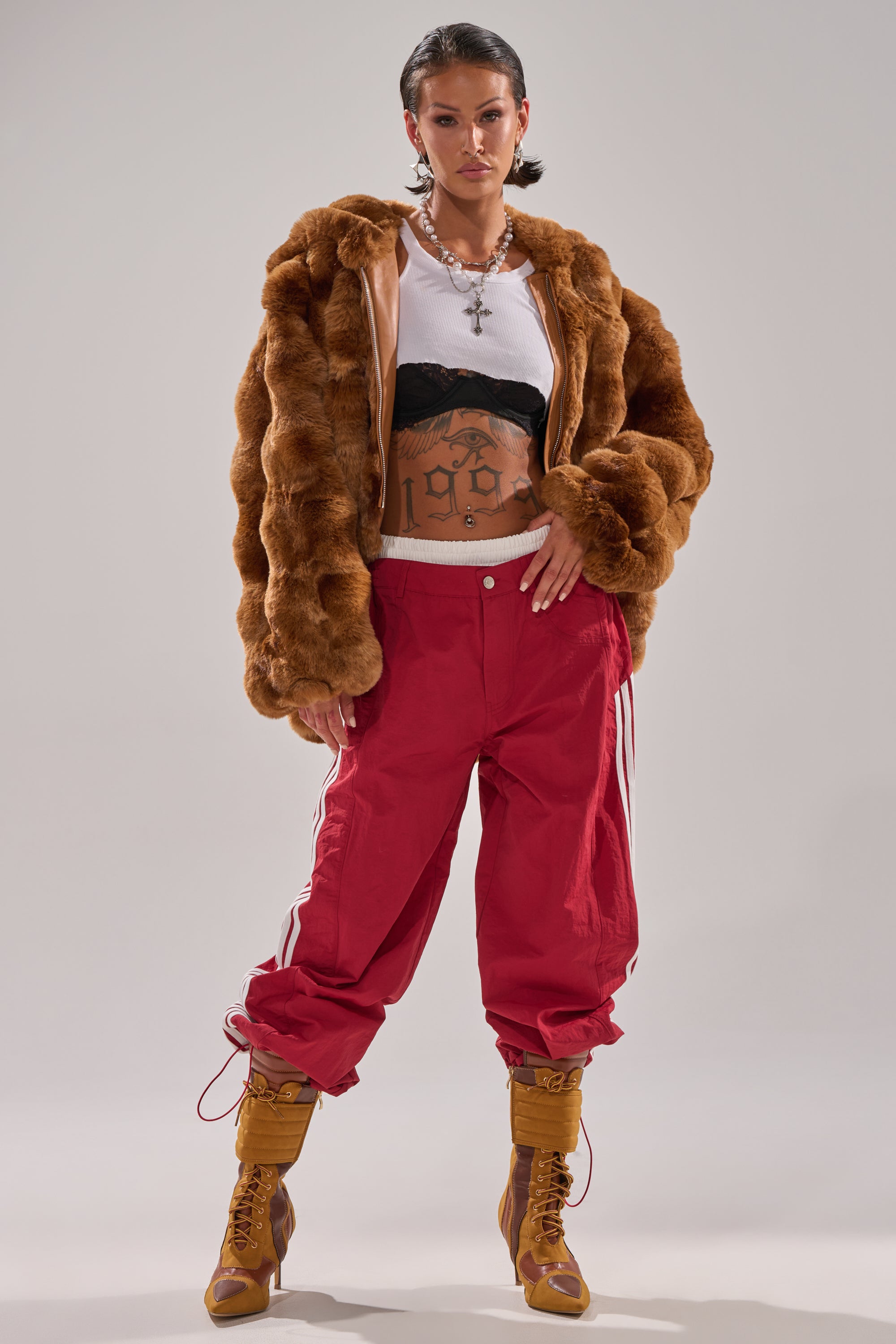 A person in a brown fur jacket, white crop top, black bralette, and DENNIS TRACK PANT IN BURGUNDY with athletic stripes stands confidently against a plain background, showcasing tattoos on their arms and torso. 4
