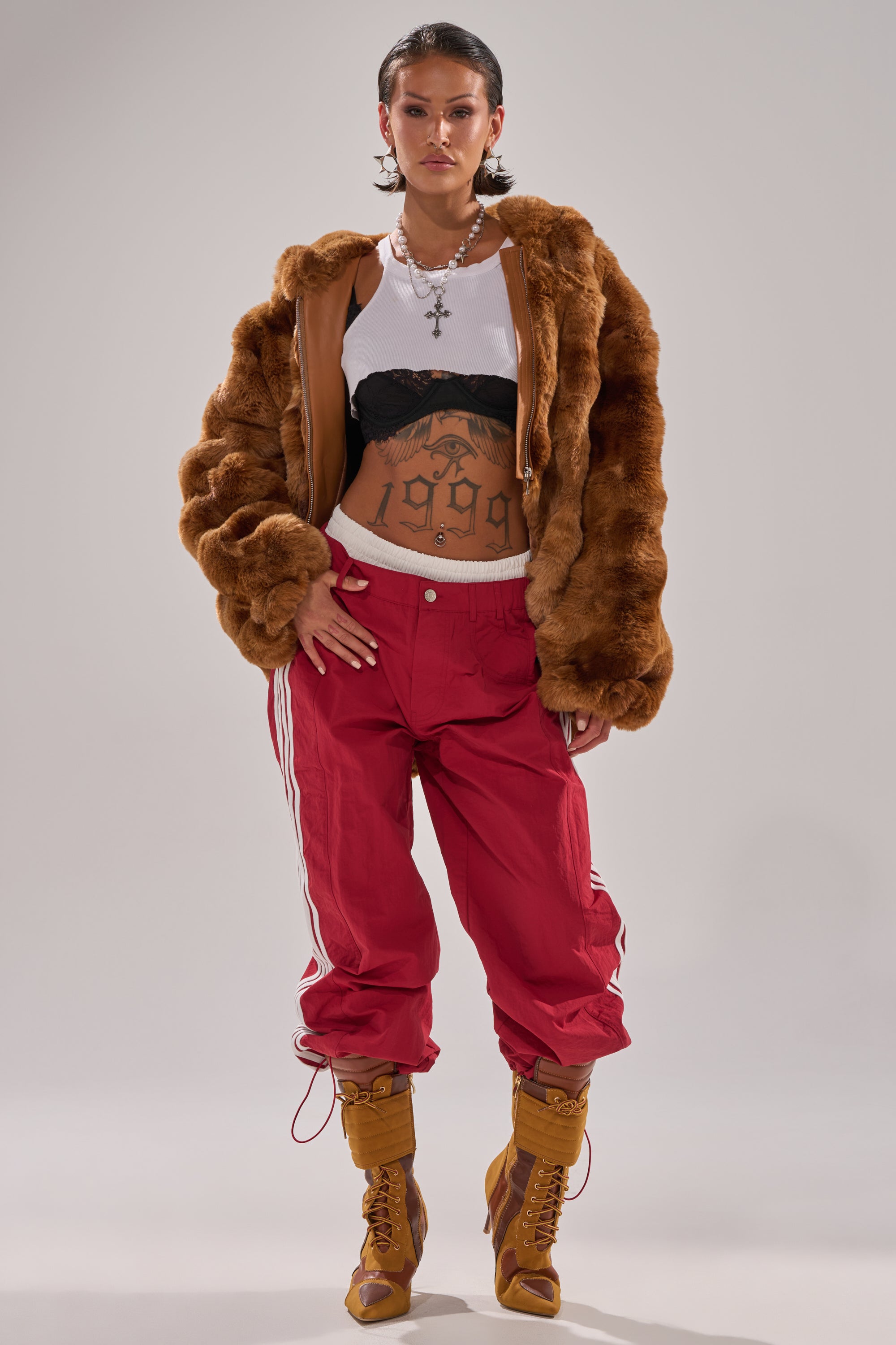 A person poses confidently in a brown fur jacket over a white crop top and black bra, paired with DENNIS TRACK PANT IN BURGUNDY, tan lace-up boots, large cross necklaces, and a "1999" tattoo across their stomach. 5