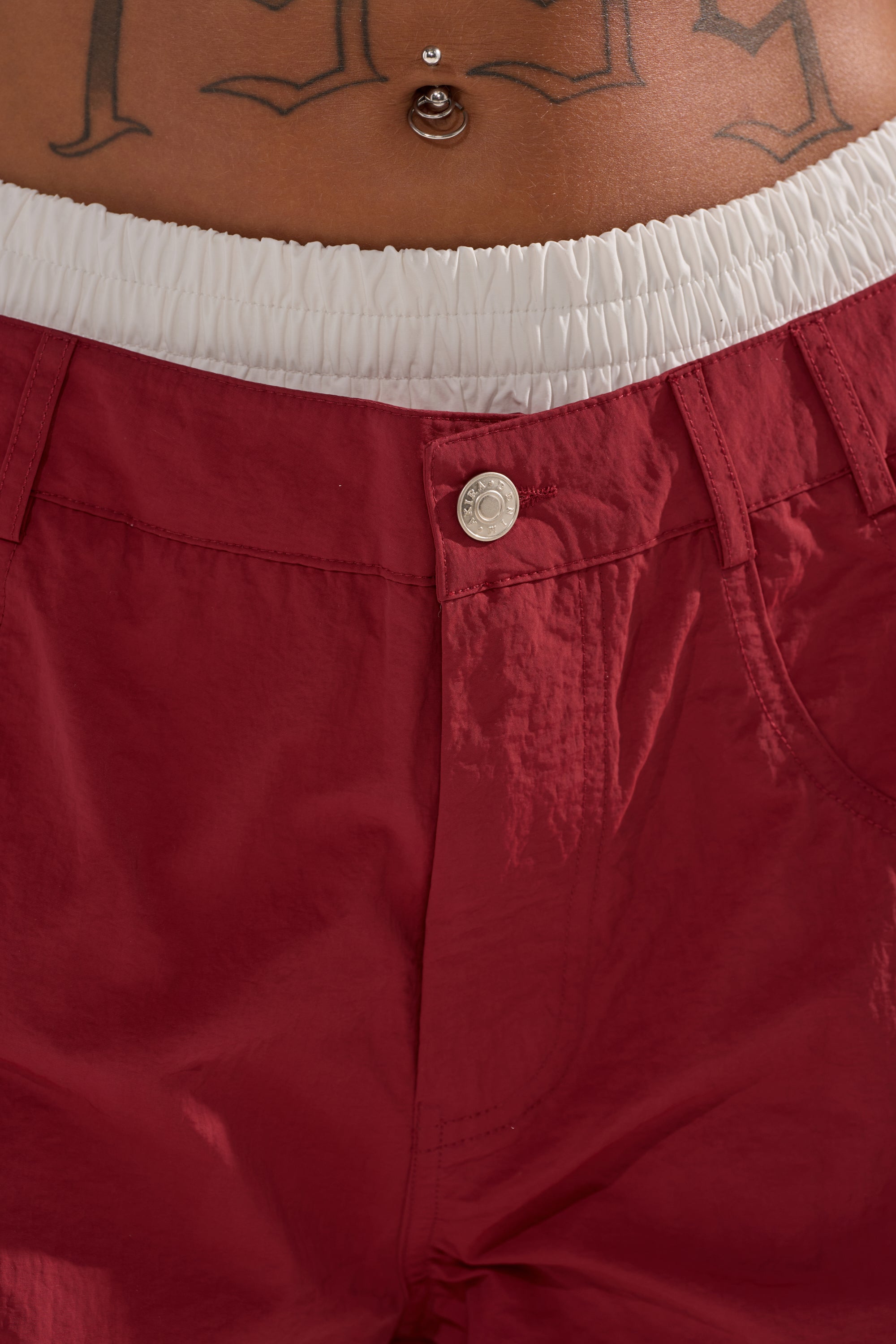 Close-up of a person’s lower torso wearing DENNIS TRACK PANT IN BURGUNDY featuring a white elastic waistband. The high-rise pants reveal a belly button piercing and a “1995” tattoo above the waistband. 6