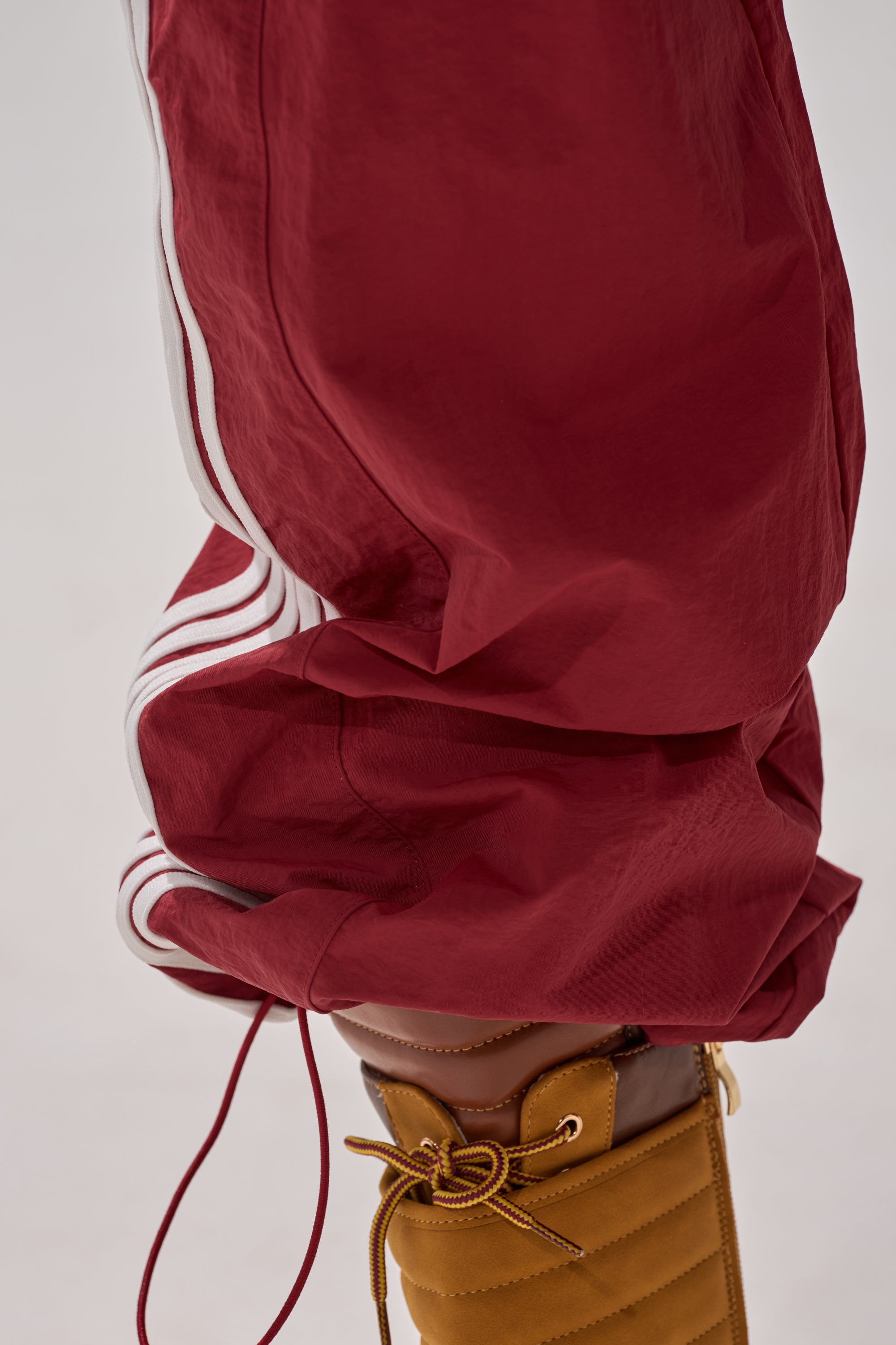 A close-up of a person wearing the DENNIS TRACK PANT IN BURGUNDY with athletic stripes, paired with tan quilted lace-up boots, set against a plain light background. 7