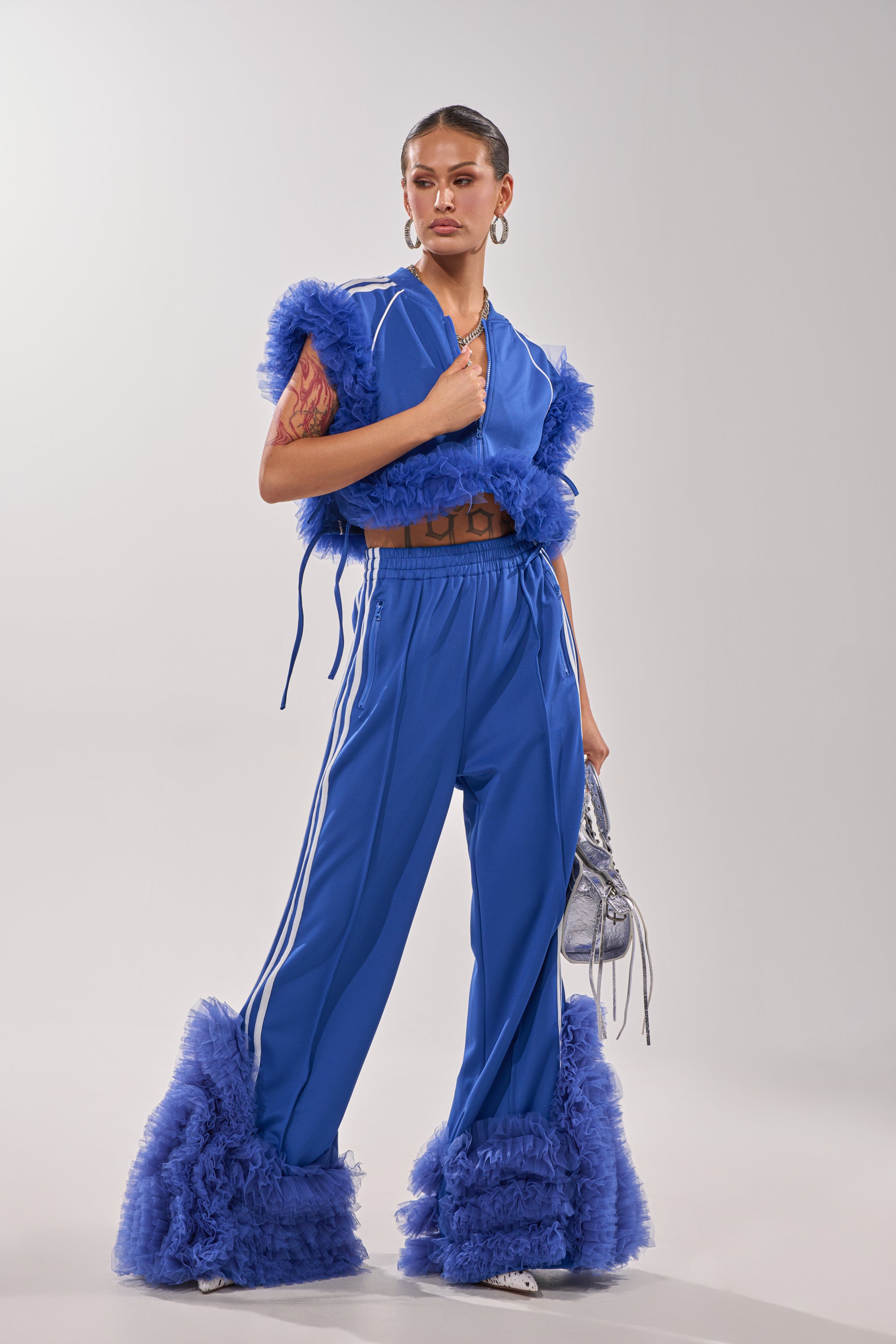 A woman poses in the MODERN JAM SWEATSHIRT WITH TULLE IN ROYAL BLUE, paired with matching high-waisted pants. She accessorizes with a small silver bag and hoop earrings, her hair slicked back for a sleek finish. 9