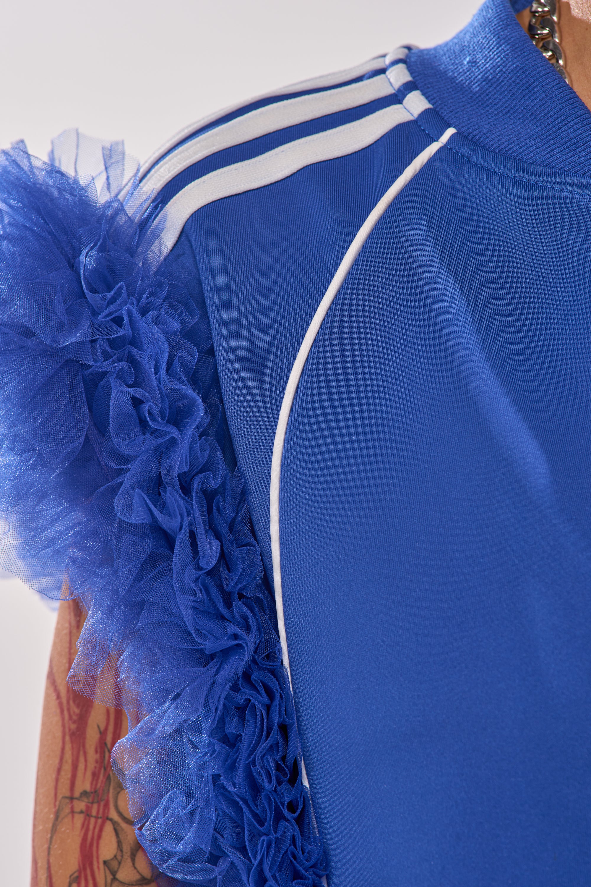 A close-up of the MODERN JAM SWEATSHIRT WITH TULLE IN ROYAL BLUE shows white shoulder stripes, ruffled tulle sleeves, and a tattooed arm, highlighting this standout athleisure piece. 10