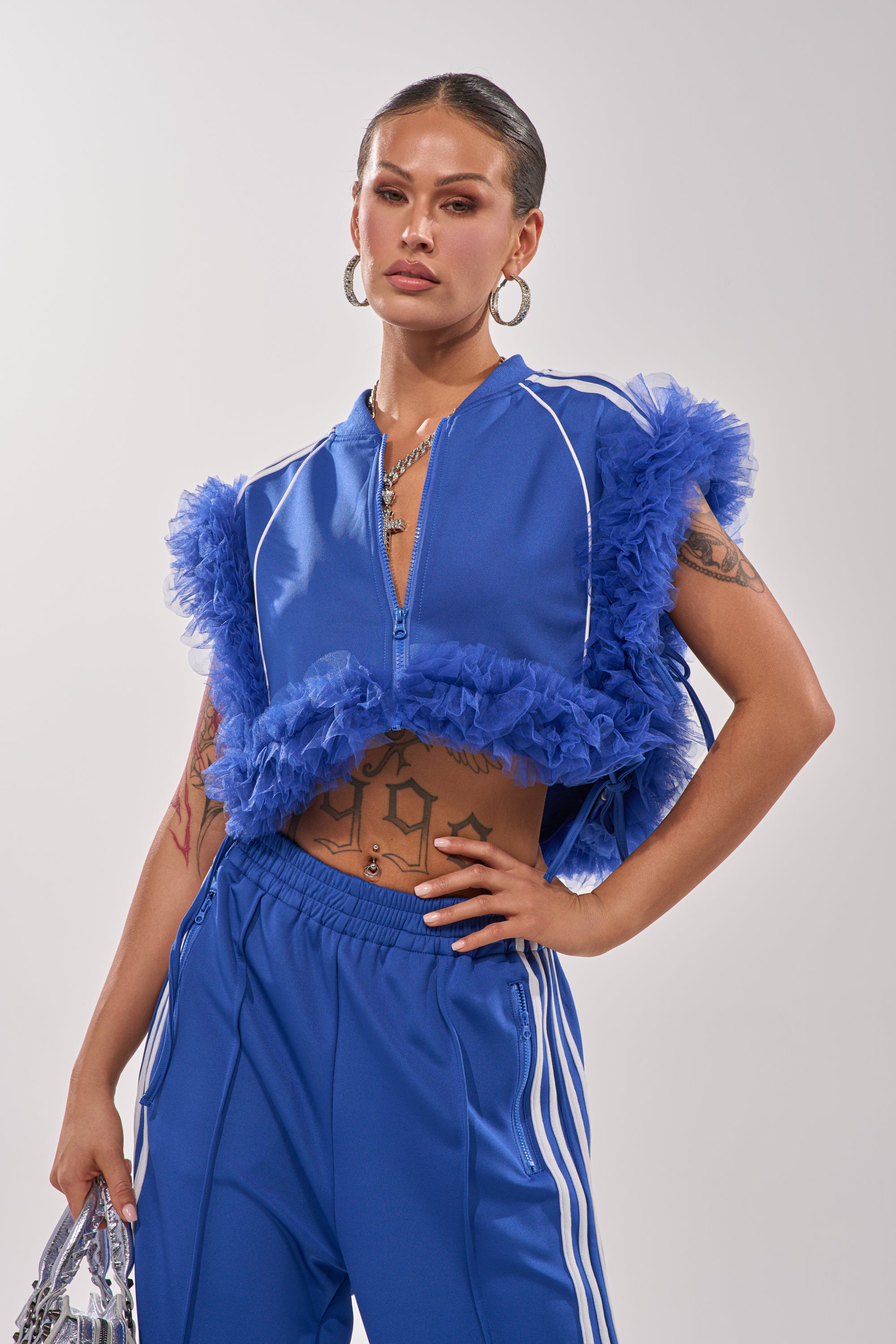 A woman poses confidently in the MODERN JAM SWEATSHIRT WITH TULLE IN ROYAL BLUE, styled with hoop earrings and a shiny silver accessory. Her slicked-back hair and visible tattoos enhance the bold look against a plain background. 2