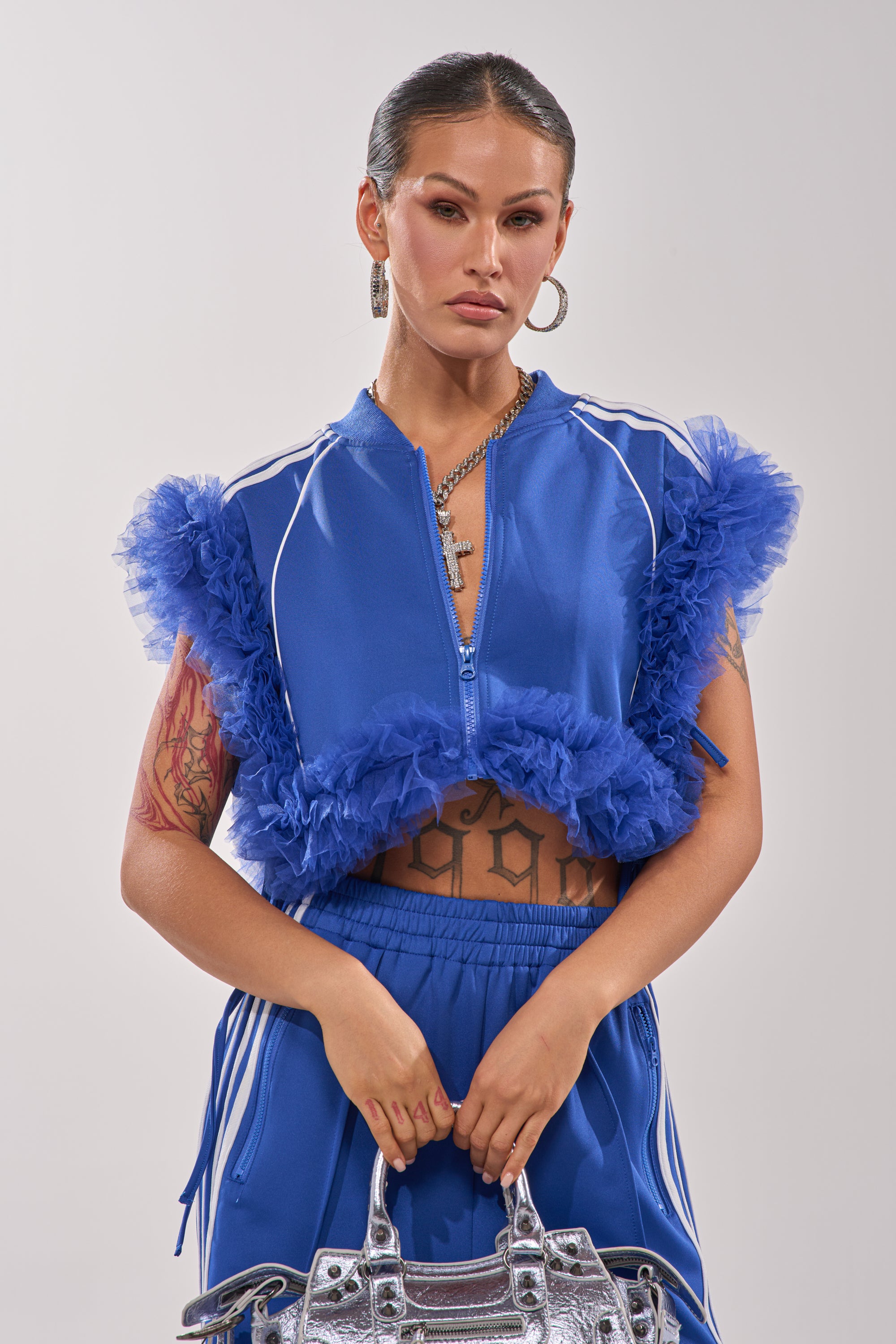 A woman with slicked-back hair and visible arm and torso tattoos wears the MODERN JAM SWEATSHIRT WITH TULLE IN ROYAL BLUE, matching pants, hoop earrings, layered necklaces, and carries a silver handbag. 3