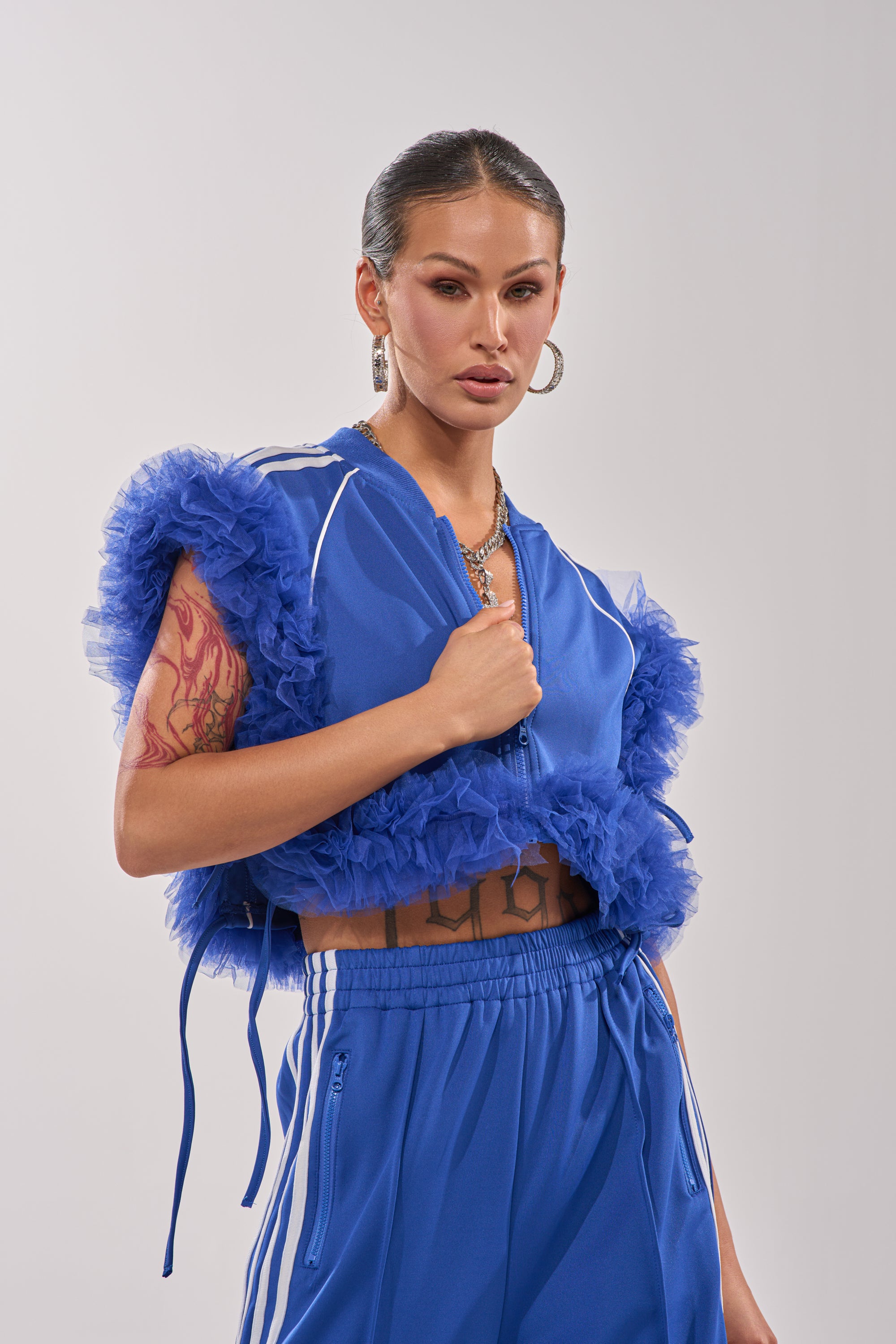 A woman stands confidently in the MODERN JAM SWEATSHIRT WITH TULLE IN ROYAL BLUE and matching pants, accessorized with hoop earrings, a necklace, and tattoos on her arms and midriff against a plain light background. 4