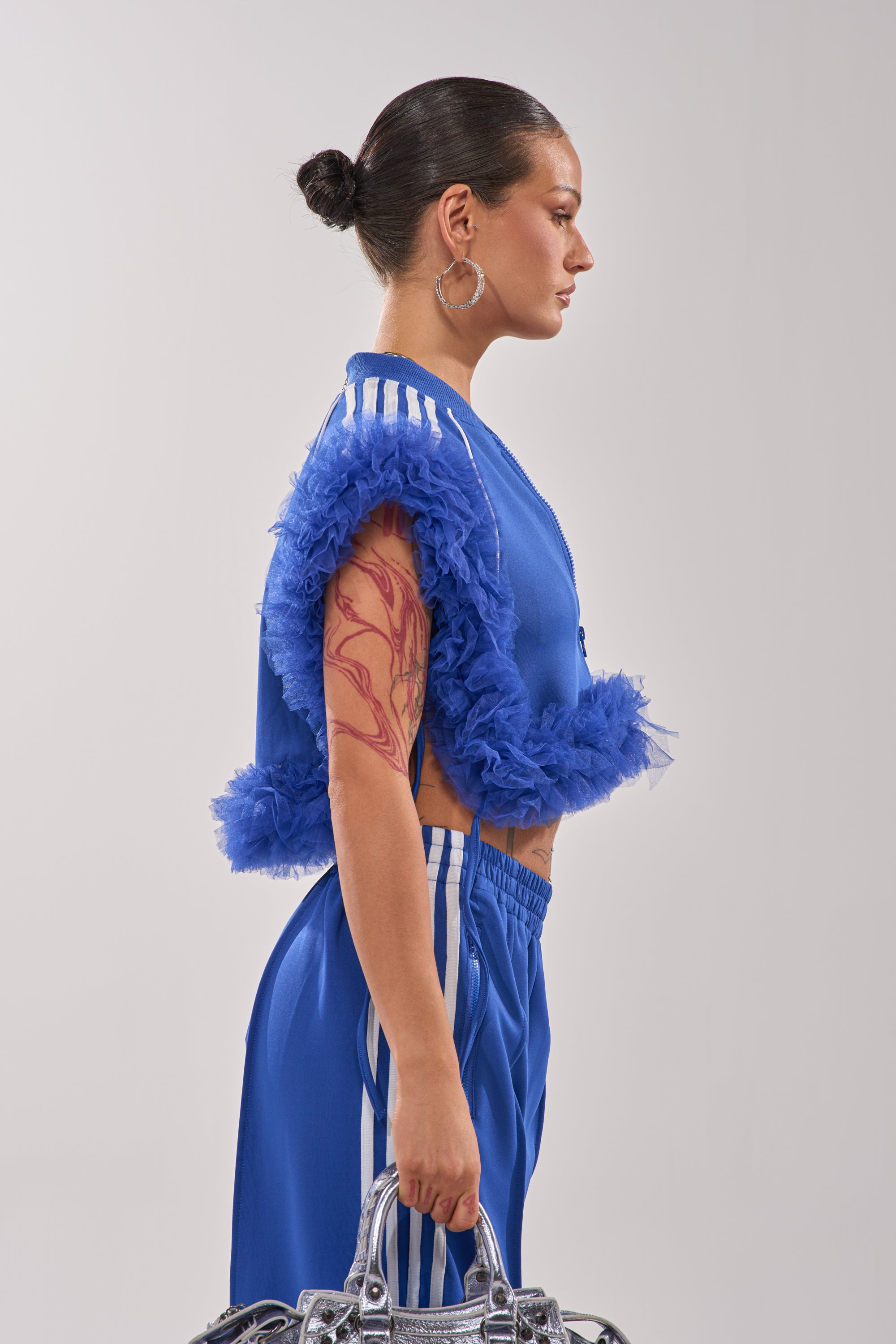 A woman with a bun and large arm tattoo, shown in profile against a plain background, wears the MODERN JAM SWEATSHIRT WITH TULLE IN ROYAL BLUE and holds a metallic silver bag. 5