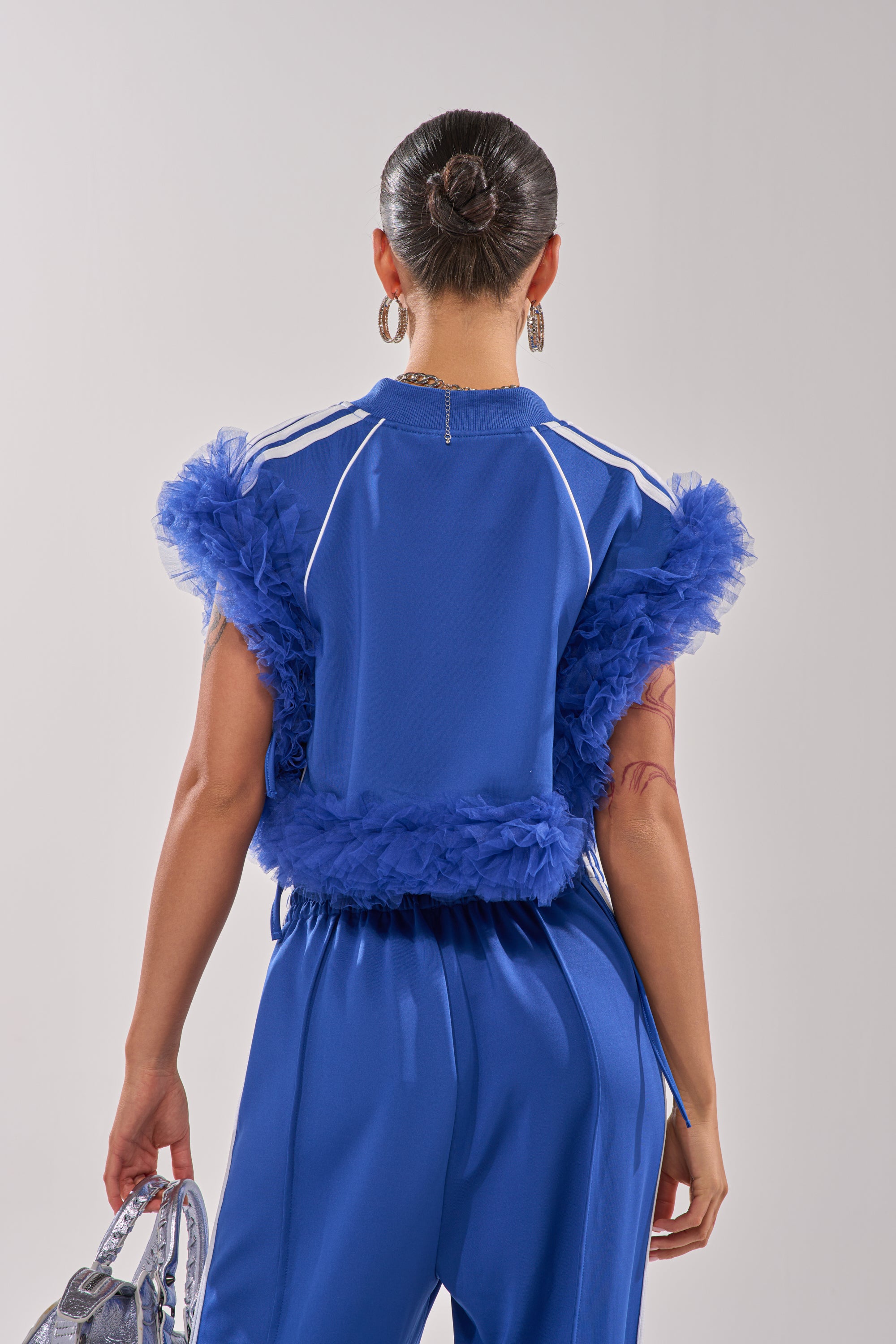A person with a neat bun, seen from behind against a plain light background, wears the MODERN JAM SWEATSHIRT WITH TULLE IN ROYAL BLUE featuring dramatic sleeves and waistband, and holds a silver bag. 6