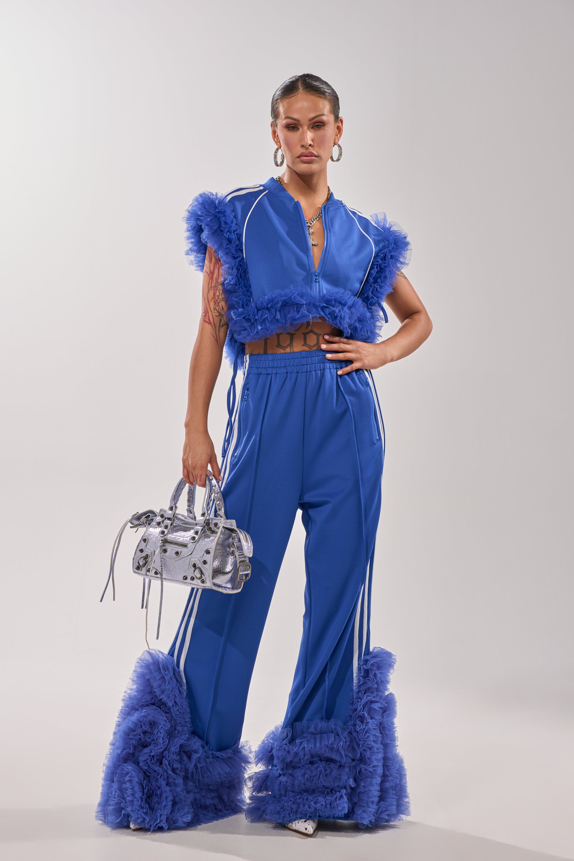 A woman poses confidently in the MODERN JAM SWEATSHIRT WITH TULLE IN ROYAL BLUE, paired with flared pants. She carries a small silver handbag, wears hoop earrings, and has her hair slicked back. 7
