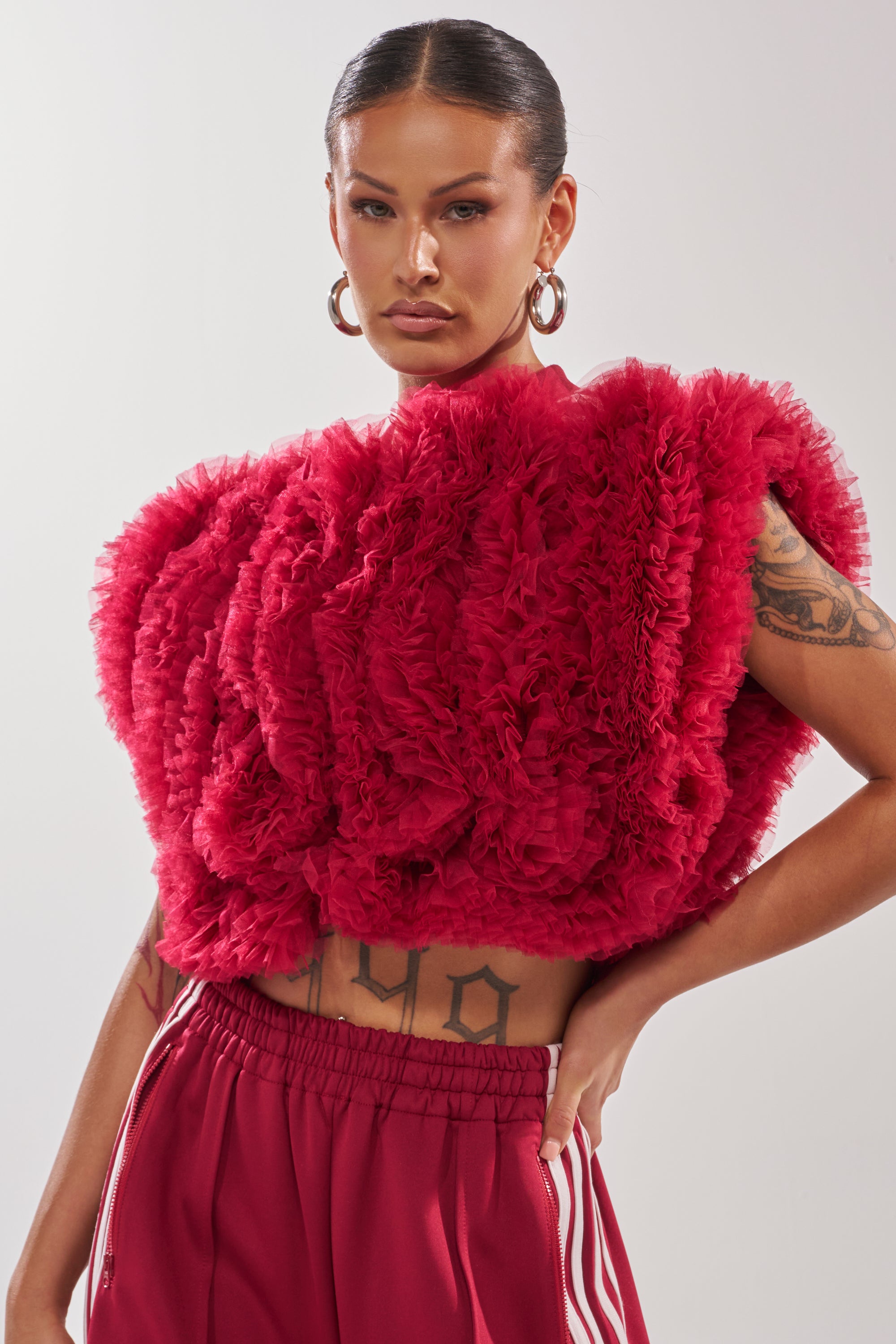 A person with slicked-back hair and tattoos poses confidently against a plain background, wearing large hoop earrings and the MODERN JAM SWEATSHIRT WITH TULLE IN BURGUNDY, paired with bold red track pants featuring white stripes. 0