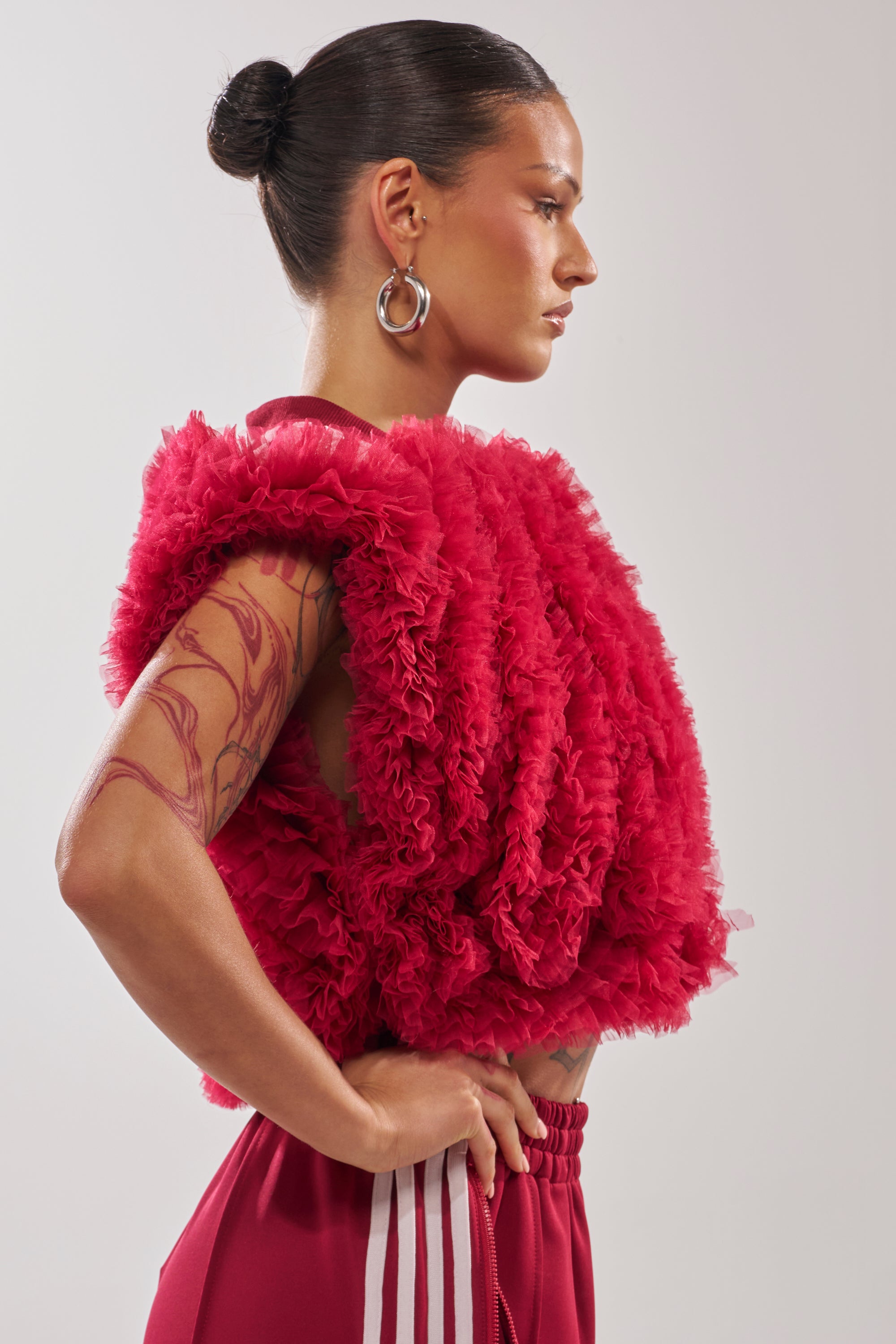 A woman with dark hair in a bun, large hoop earrings, and an arm tattoo stands in profile with her hands on her hips, wearing red striped pants and the MODERN JAM SWEATSHIRT WITH TULLE IN BURGUNDY. 2