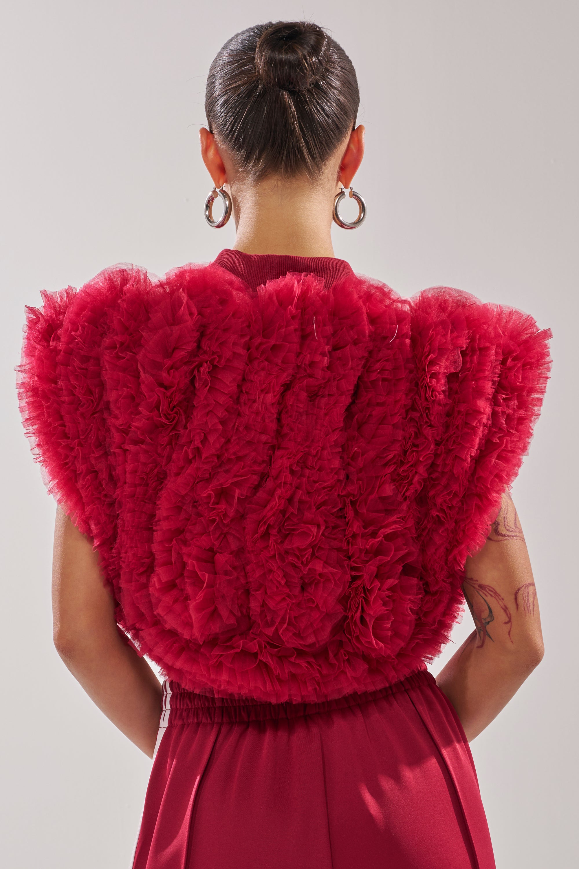 A person with dark hair in a bun, wearing large hoop earrings and the MODERN JAM SWEATSHIRT WITH TULLE IN BURGUNDY, is seen from behind with a tattoo on their right upper arm. 3