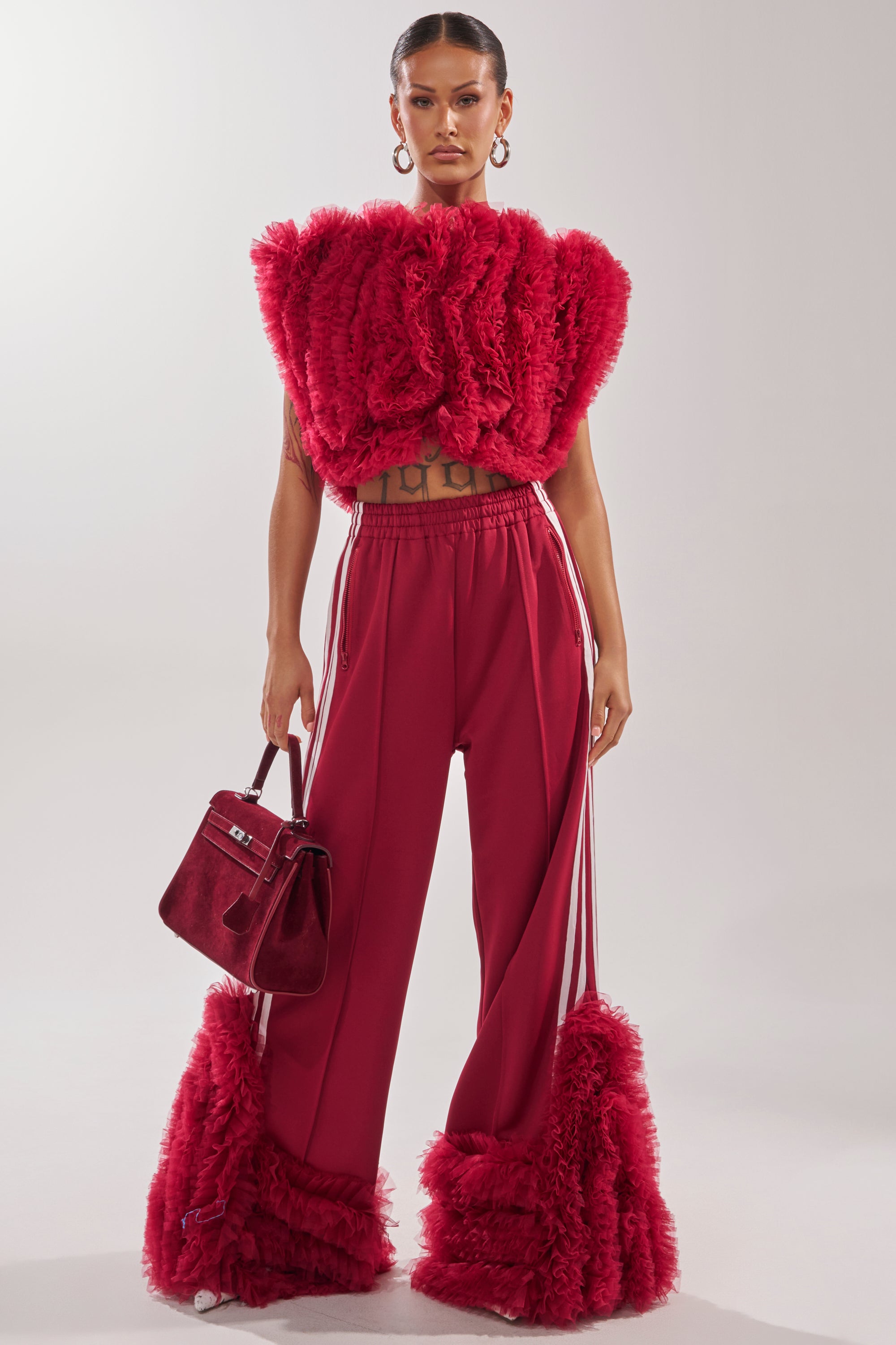 A woman faces forward in the MODERN JAM SWEATSHIRT WITH TULLE IN BURGUNDY paired with wide-legged ruffled pants. She carries a red handbag, wears large hoop earrings, and has her hair pulled back tightly. 4
