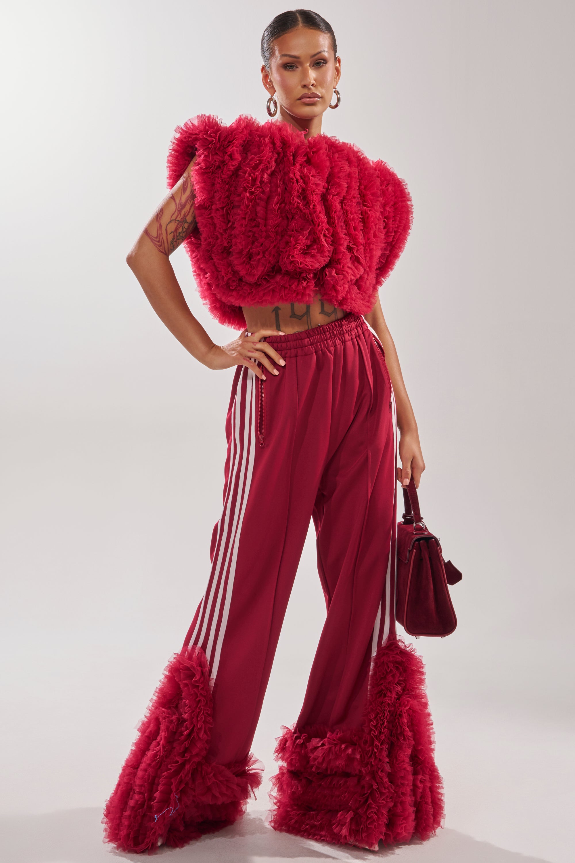 A person poses confidently in the MODERN JAM SWEATSHIRT WITH TULLE IN BURGUNDY, paired with matching pants featuring white stripes and furry ankle trim, holding a red bag against a plain background. 5