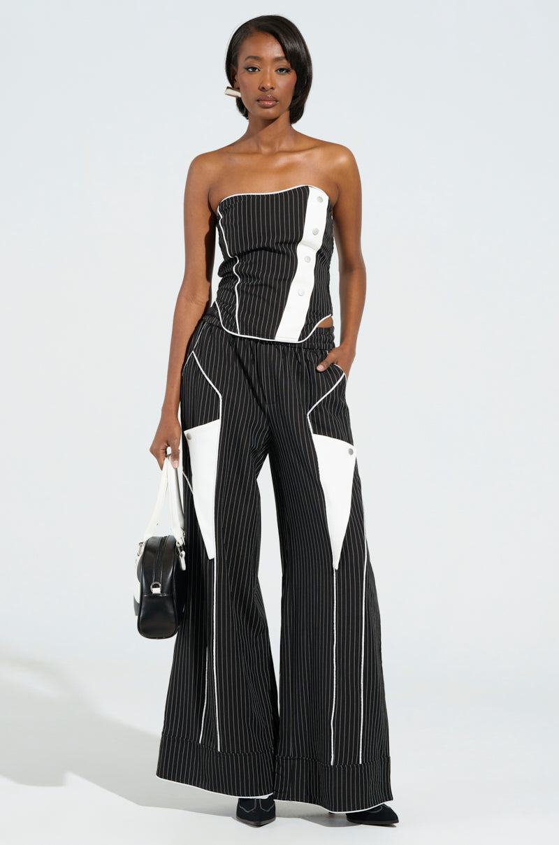 A woman stands confidently, wearing the Strictly Business Pinstripe Pant, a black and white pinstriped strapless jumpsuit with a wide-leg flared design and white detailing. She holds a black handbag, set against a plain, light background.